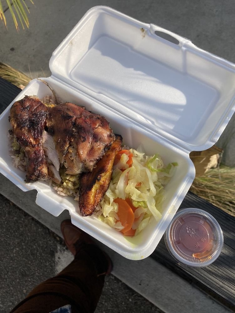 Takeout container with grilled chicken, rice, and coleslaw, on a wooden surface outside, with a small cup of juice nearby.
