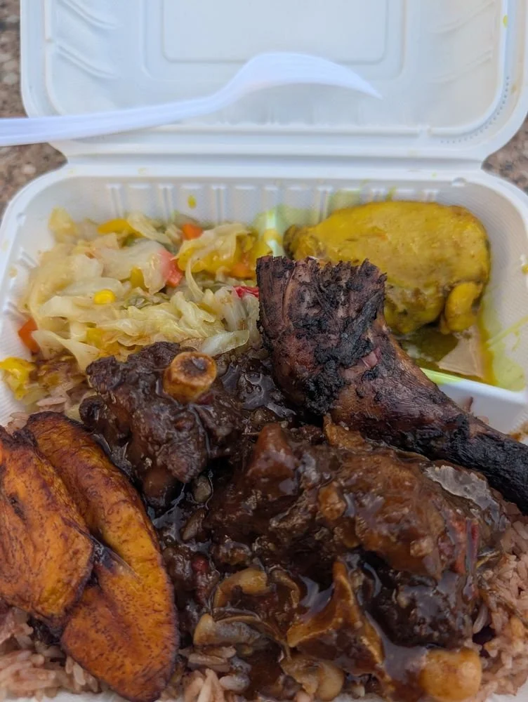 Takeout container with fried plantains, braised meat with sauce, sautéed vegetables, and a yellow curry chicken drumstick.