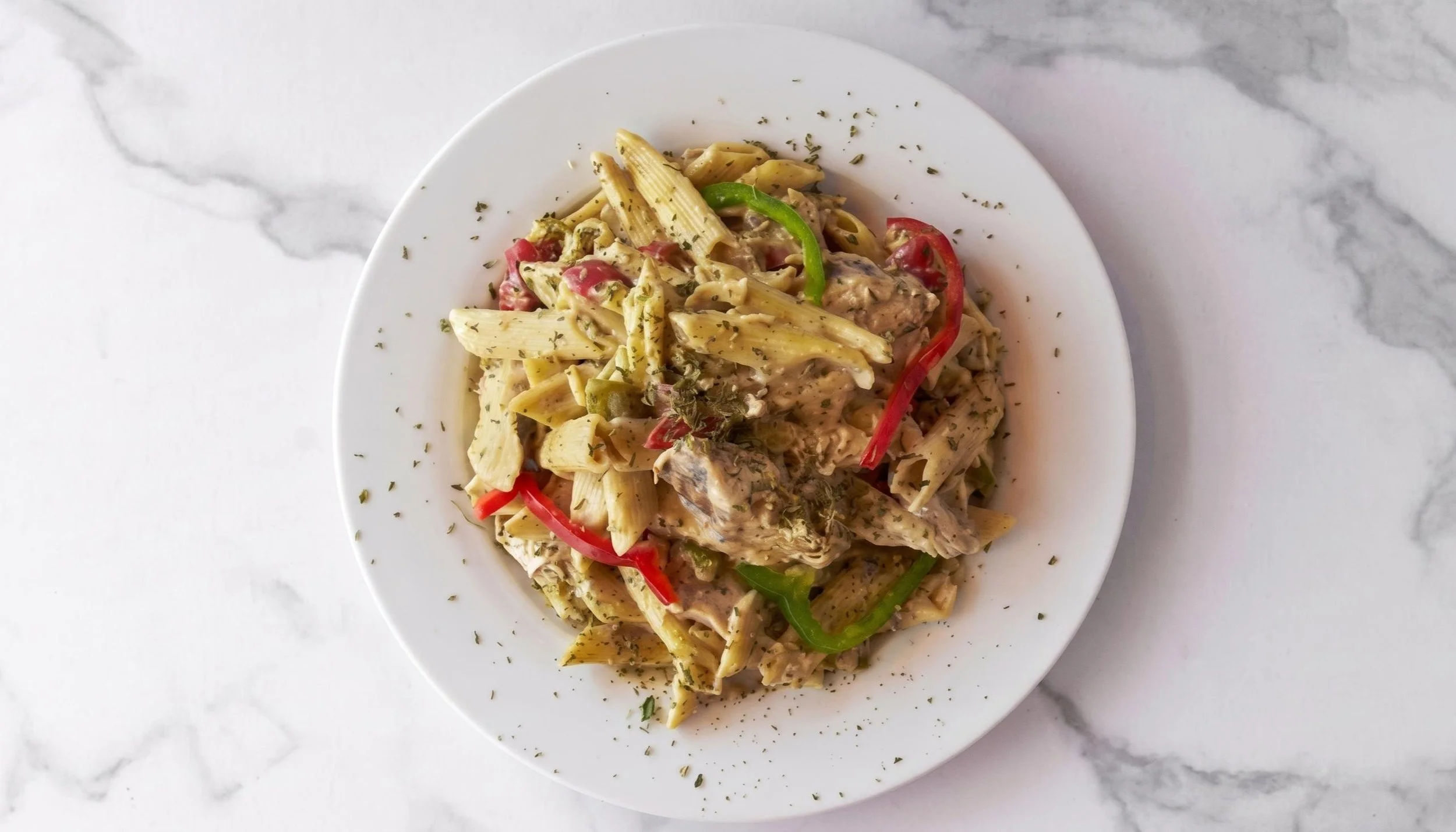 Plate of cooked penne pasta with green and red bell peppers and herbs on a marble surface.