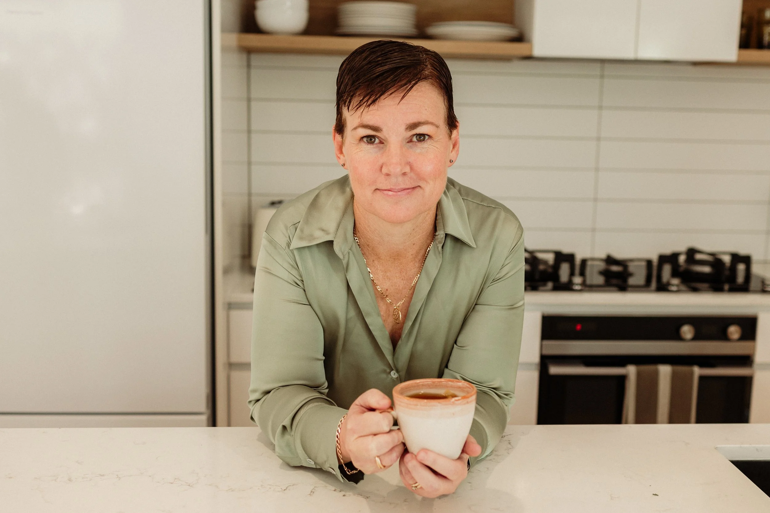 A woman with short brown hair, wearing a light green blouse, sits at a kitchen counter holding a white mug with a brown drink inside. The kitchen has white cabinets, a stove, and shelves with plates and bowls in the background.