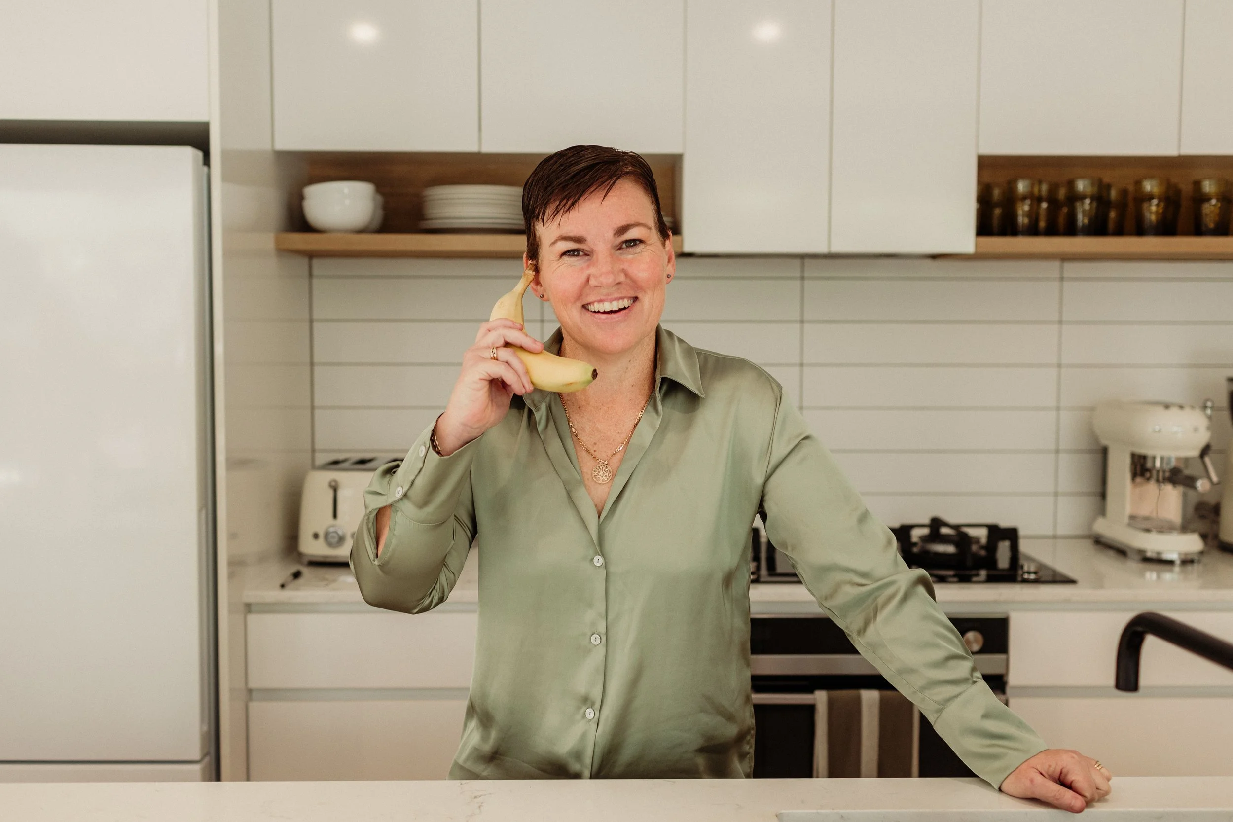 Sarah Franklin, naturopath and founder of Balanced Health Naturopathy, smiling and playfully holding a banana to her ear like a phone in a kitchen
