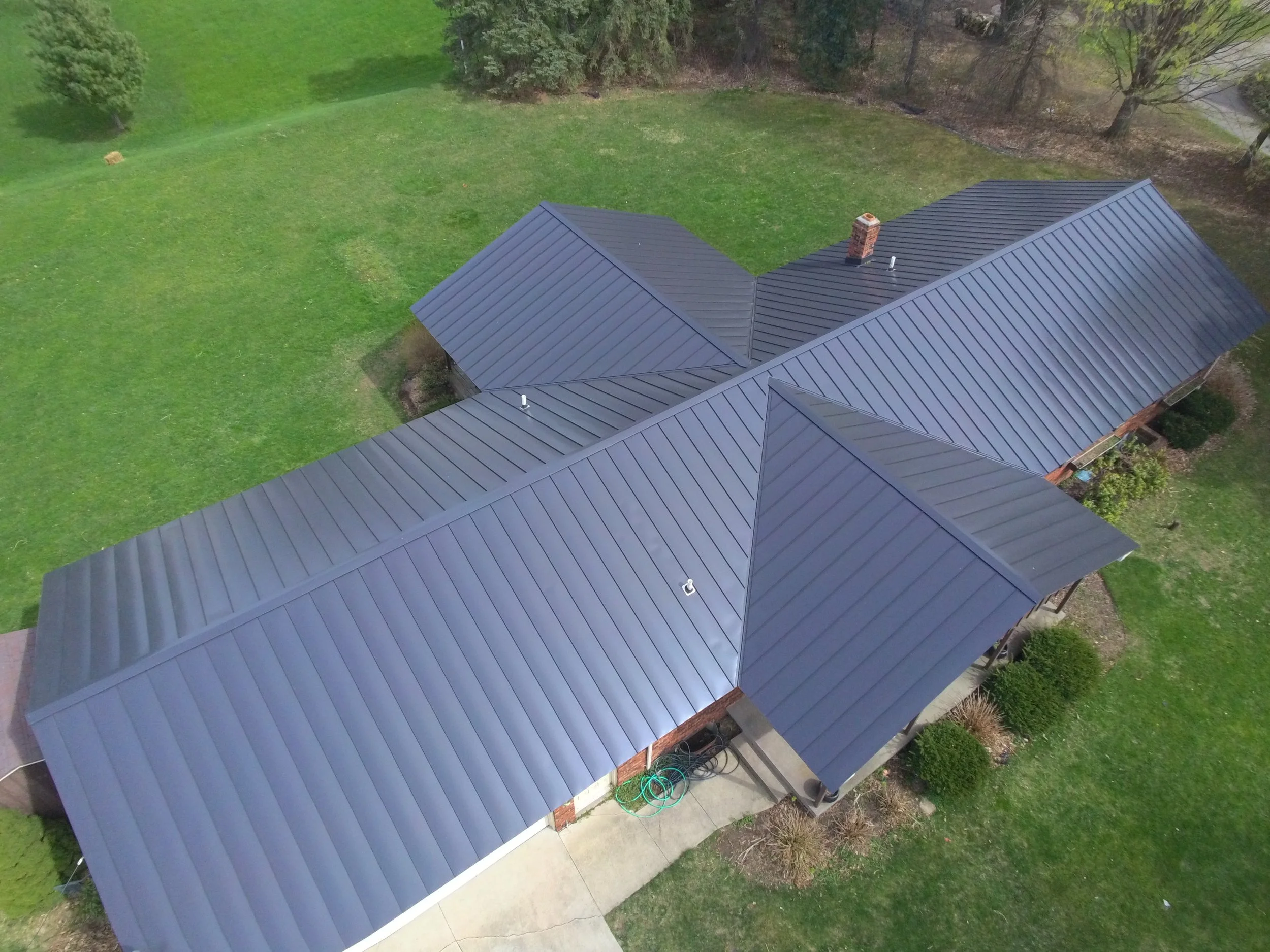 Aerial view of a house with a metal roof, surrounded by green lawn and trees.