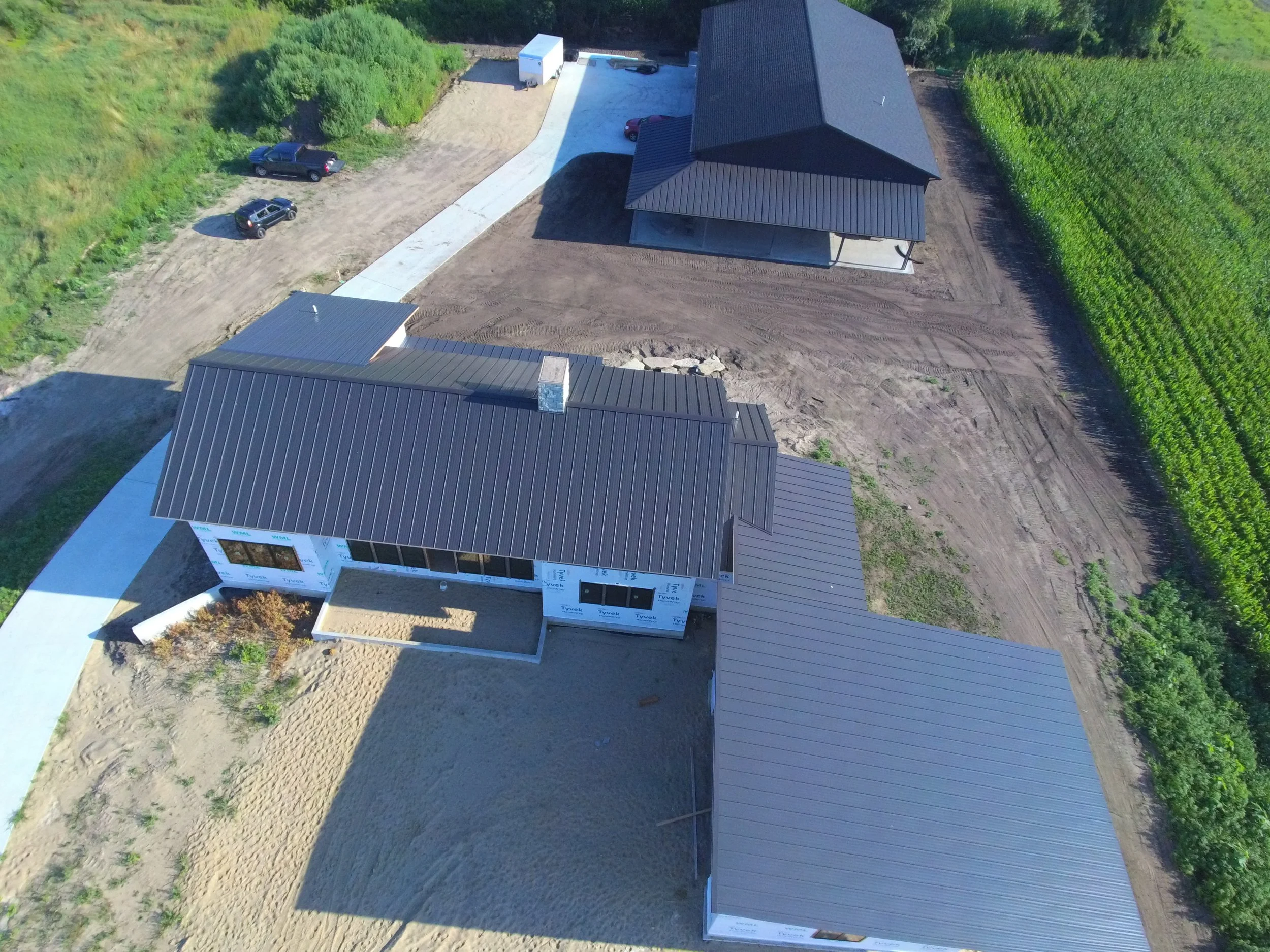Aerial view of a house under construction with new roofing being installed, surrounded by a dirt yard, parked trucks, and green fields.
