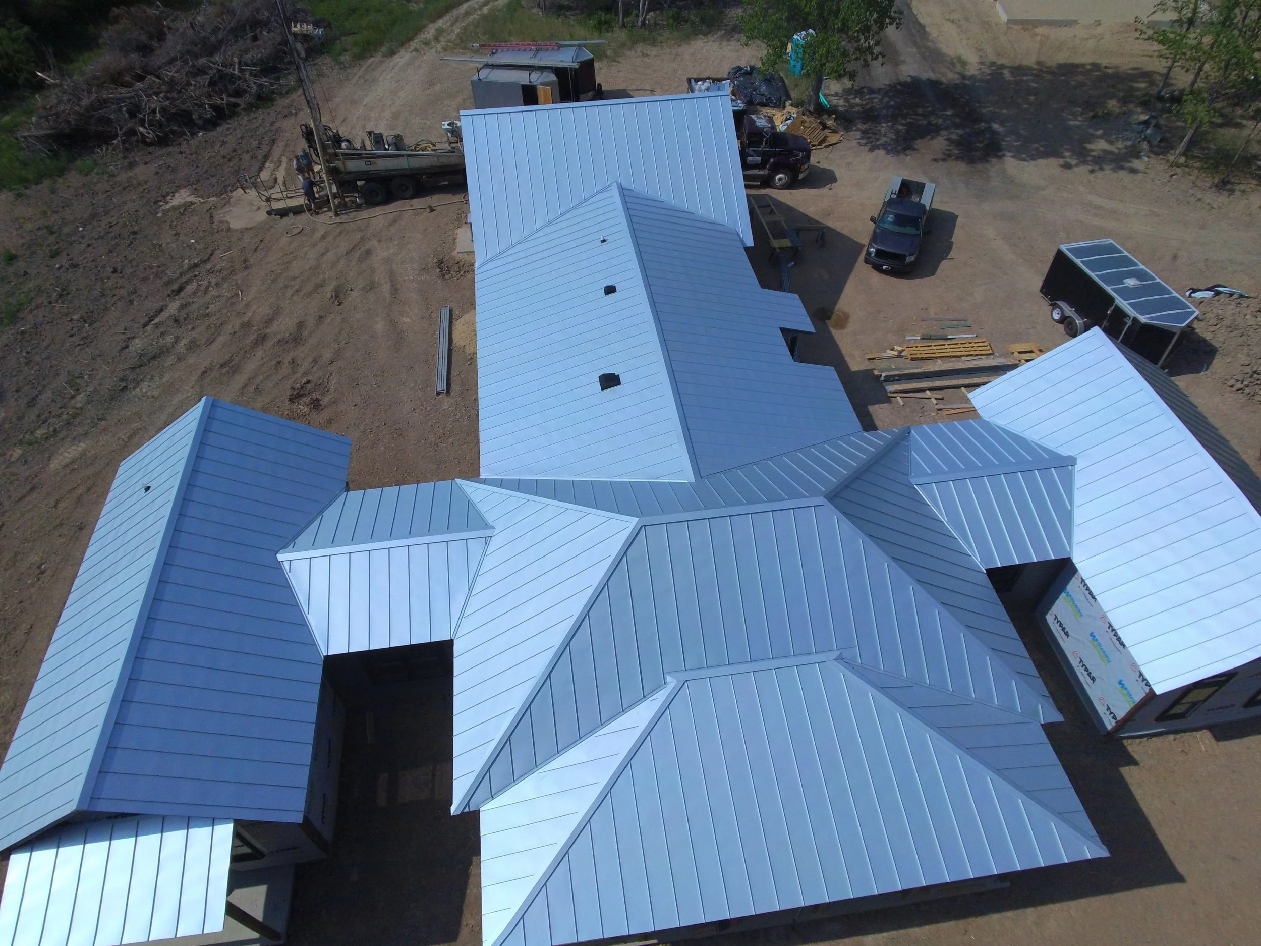 Aerial view of a construction site with a white metal roof building, construction vehicles, tools, and materials on dirt ground.