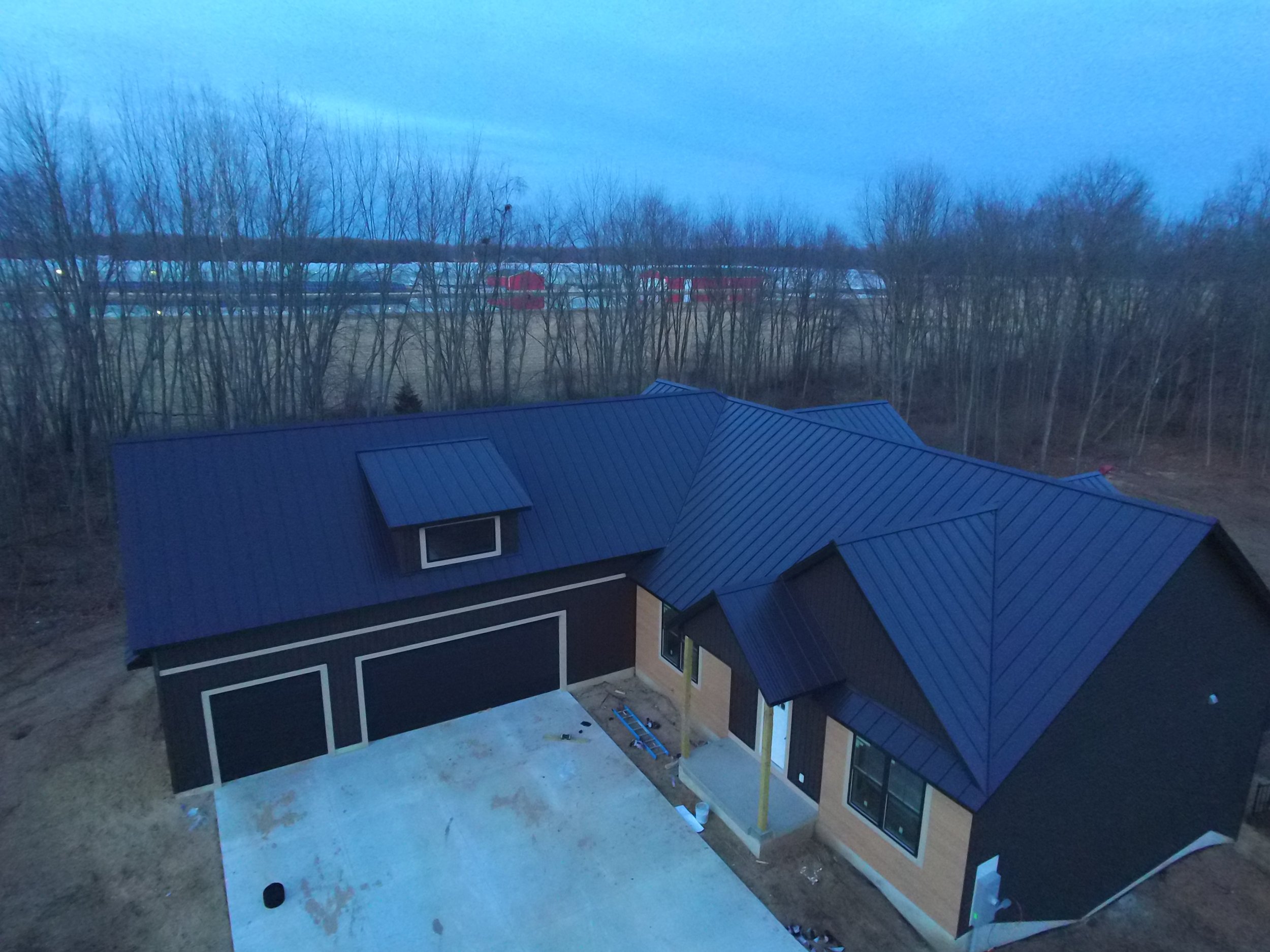 A house under construction with a black roof, wooden siding, and large windows, surrounded by a yard with a concrete driveway and trees in the background during dusk.