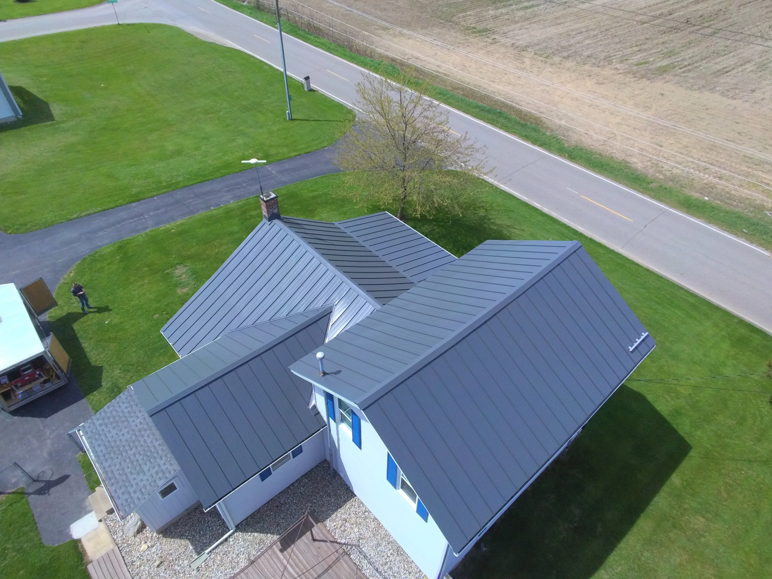 Aerial view of a house with a gray metal roof, a lawn, a tree, a driveway, and a street with utility lines.