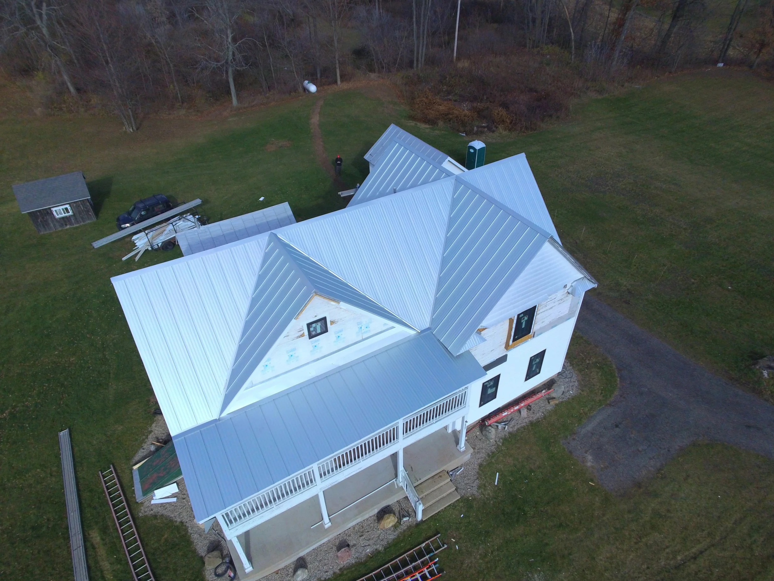 Aerial view of a house under construction with metal roof, surrounded by green lawn, trees, and construction equipment.