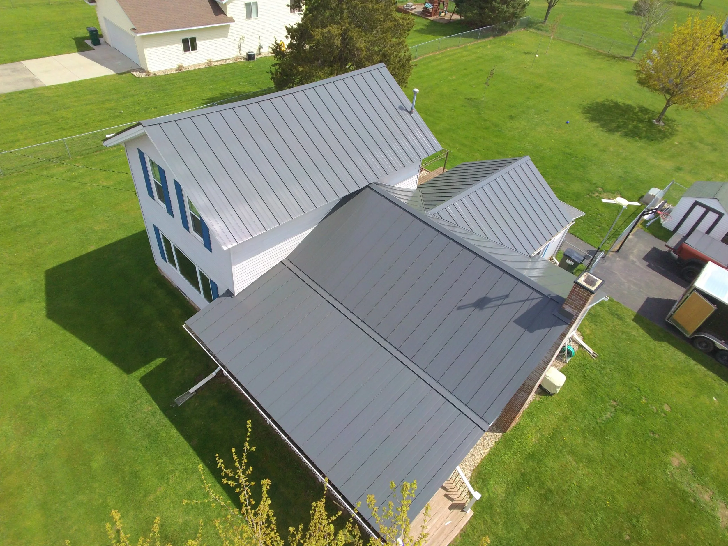 An aerial view of a residential backyard showing a two-story house with a metal roof, green lawn, trees, and surrounding fences.