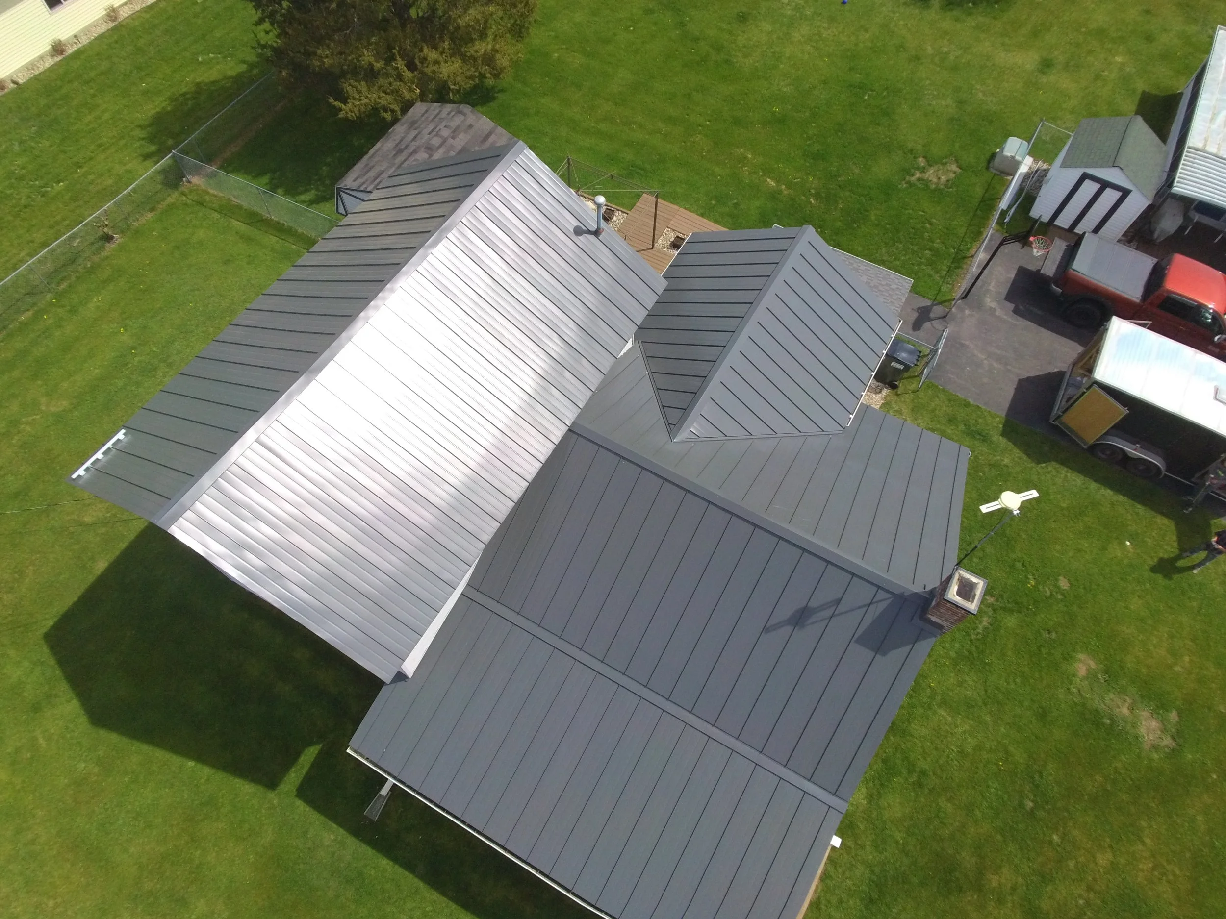 Aerial view of a backyard showing a gray metal roof covering a house, a smaller shed with a shingled roof, a lawn, and a driveway with a red vehicle parked on it.