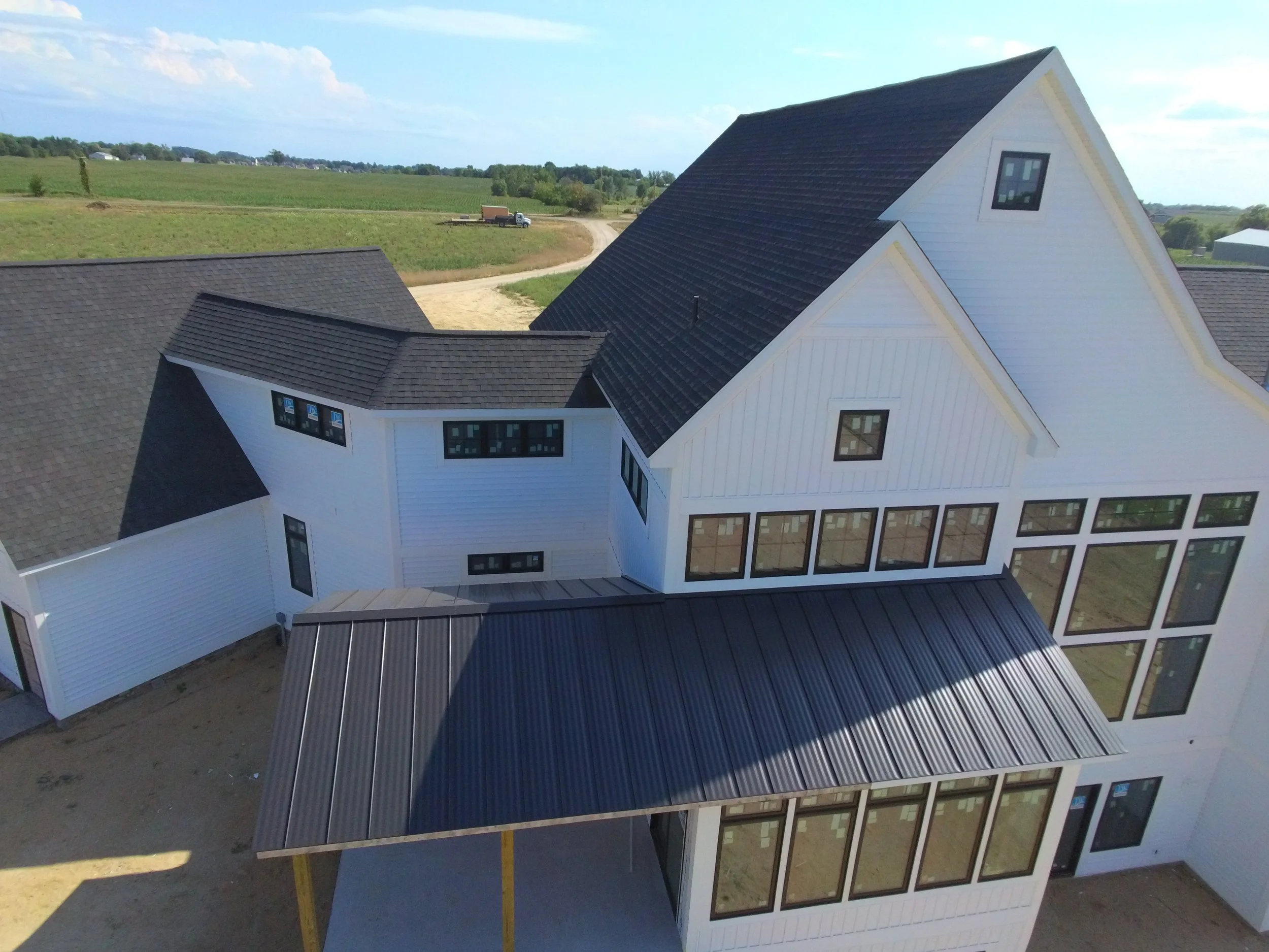 Aerial view of a modern, white residential building with multiple sections and black window frames, situated in a rural area with green fields, dirt paths, and a truck in the background.