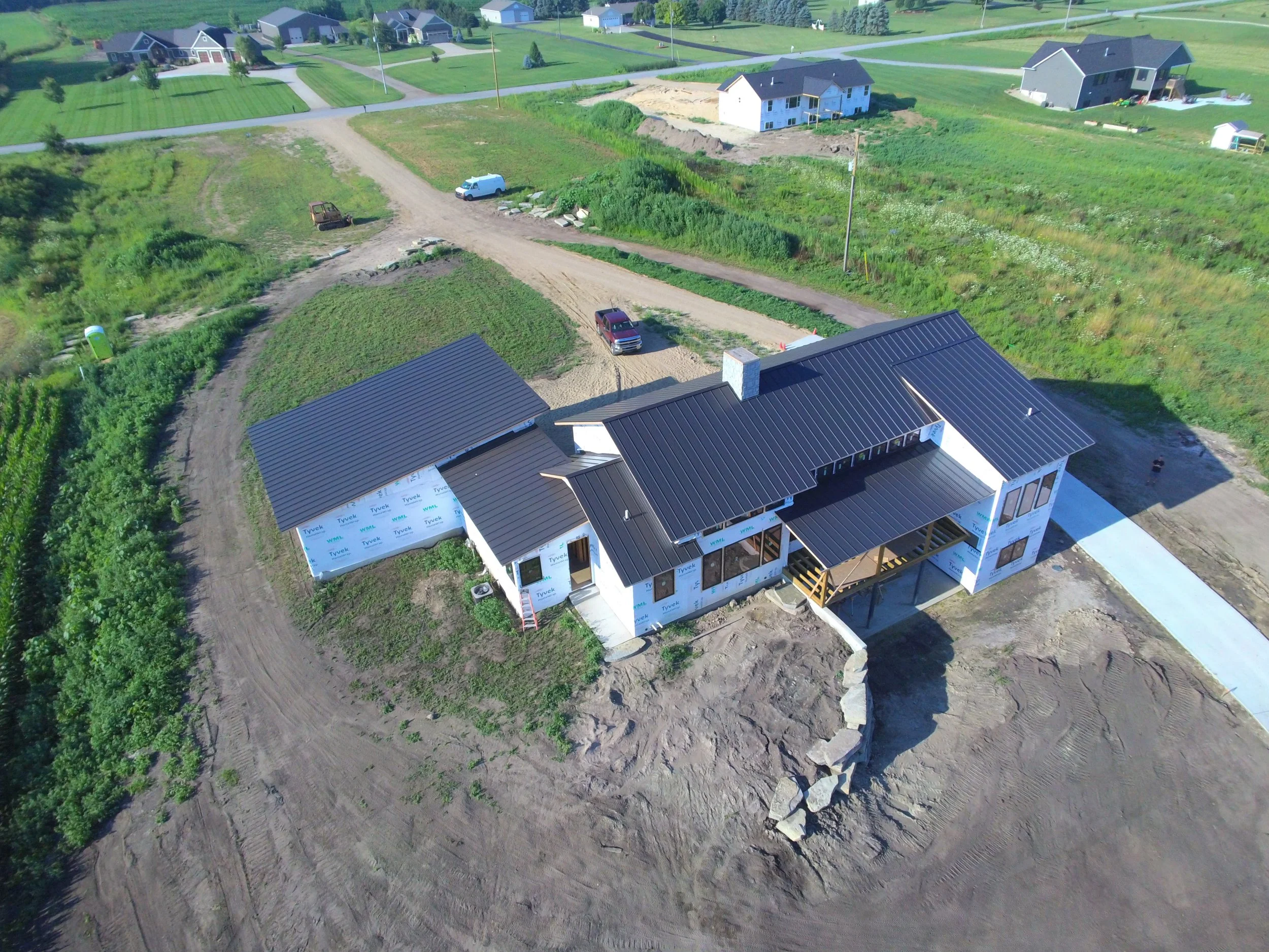 Aerial view of a house under construction with a black metal roof, surrounded by dirt and some green vegetation, with neighboring fully built houses.
