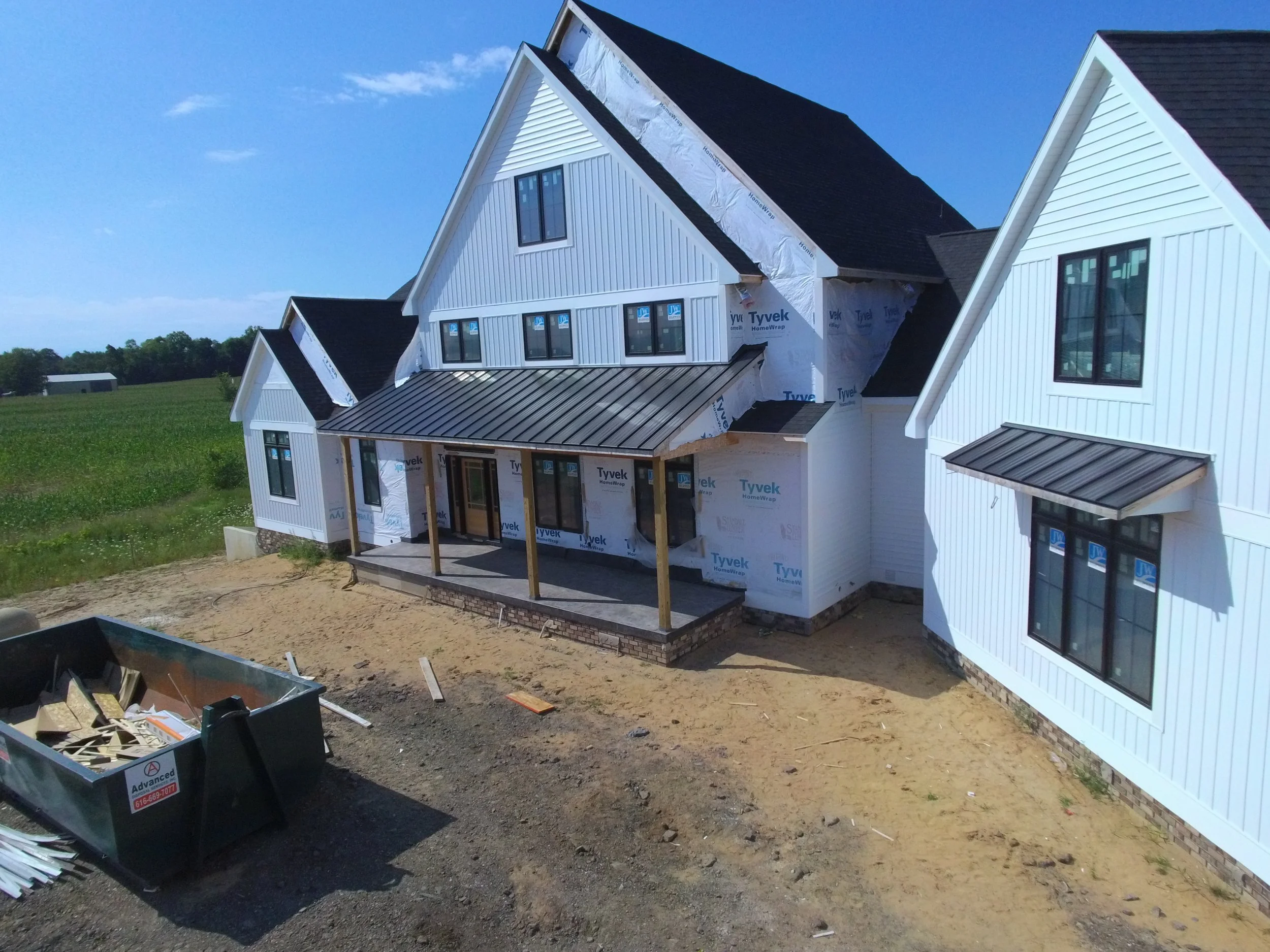 Under construction house with white siding, black roof, large windows, and a small porch, surrounded by an open field.