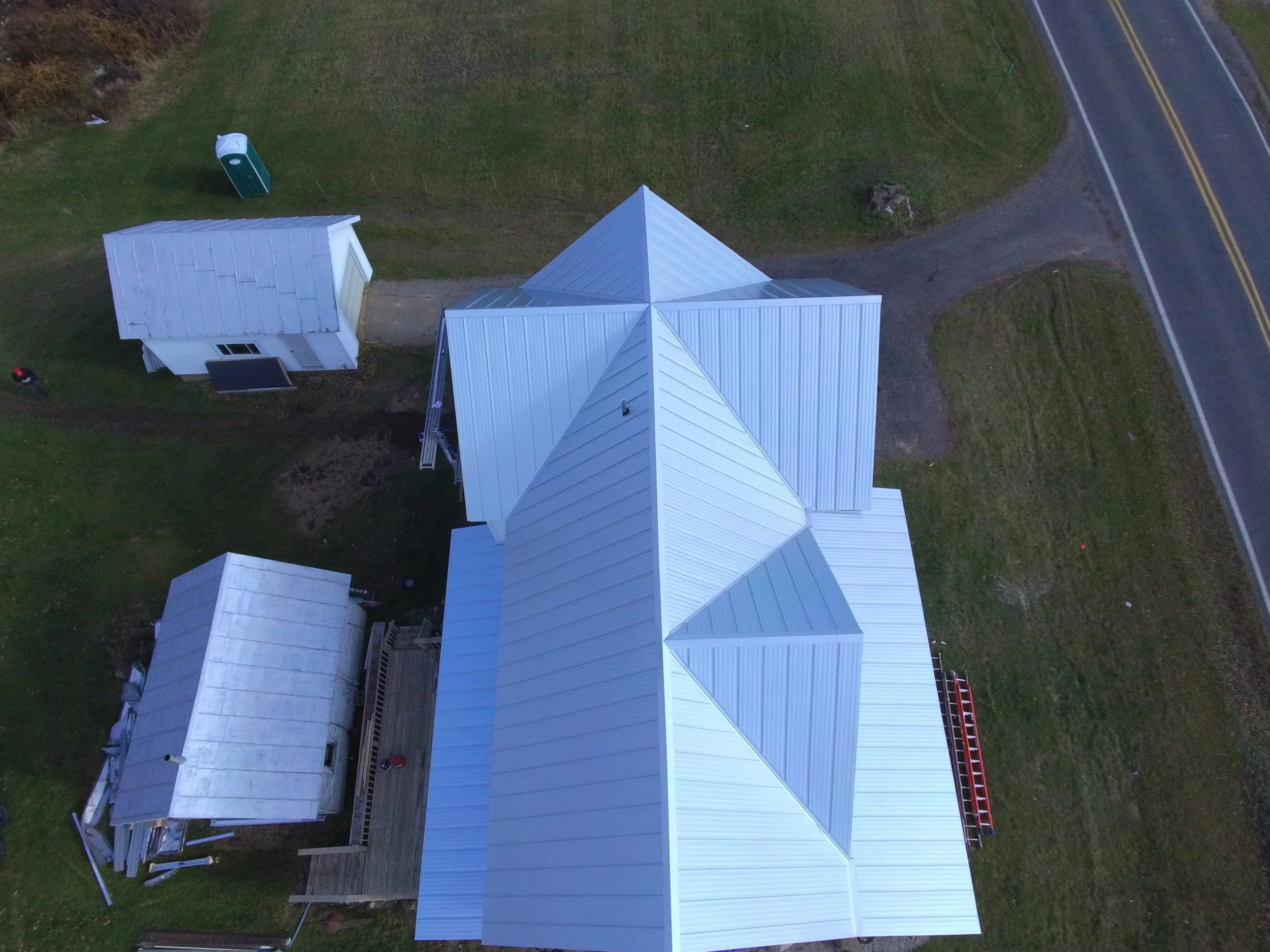 Aerial view of a white building with a pointed roof, a smaller detached structure, and an outdoor deck with some furniture, located on a grassy lot next to a paved road.