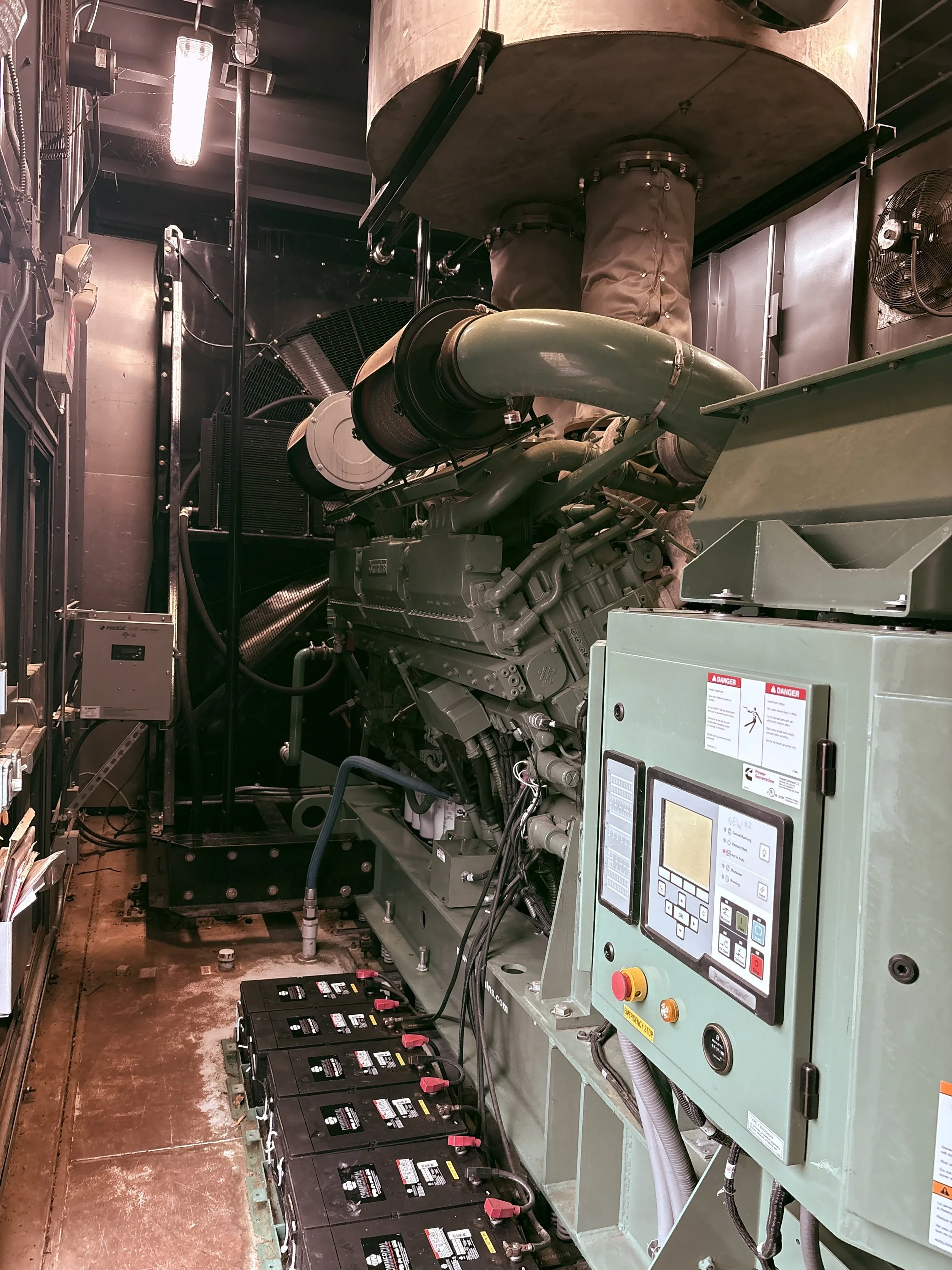 Room with large industrial generator, numerous black batteries lined up on the floor, and control panel with buttons and display.