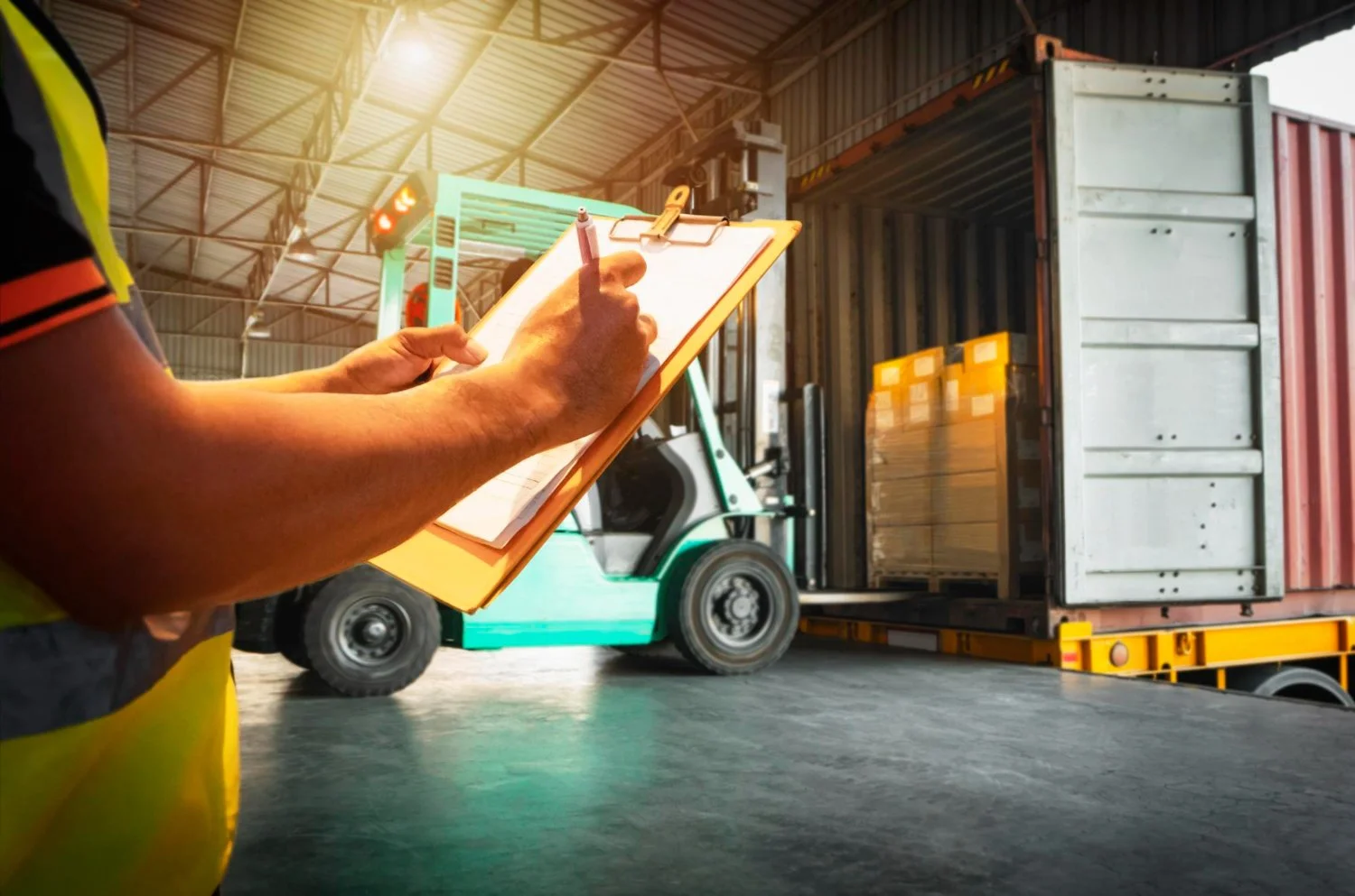 Worker on a forklift inspects cargo container being loaded in a warehouse, writing on a clipboard.