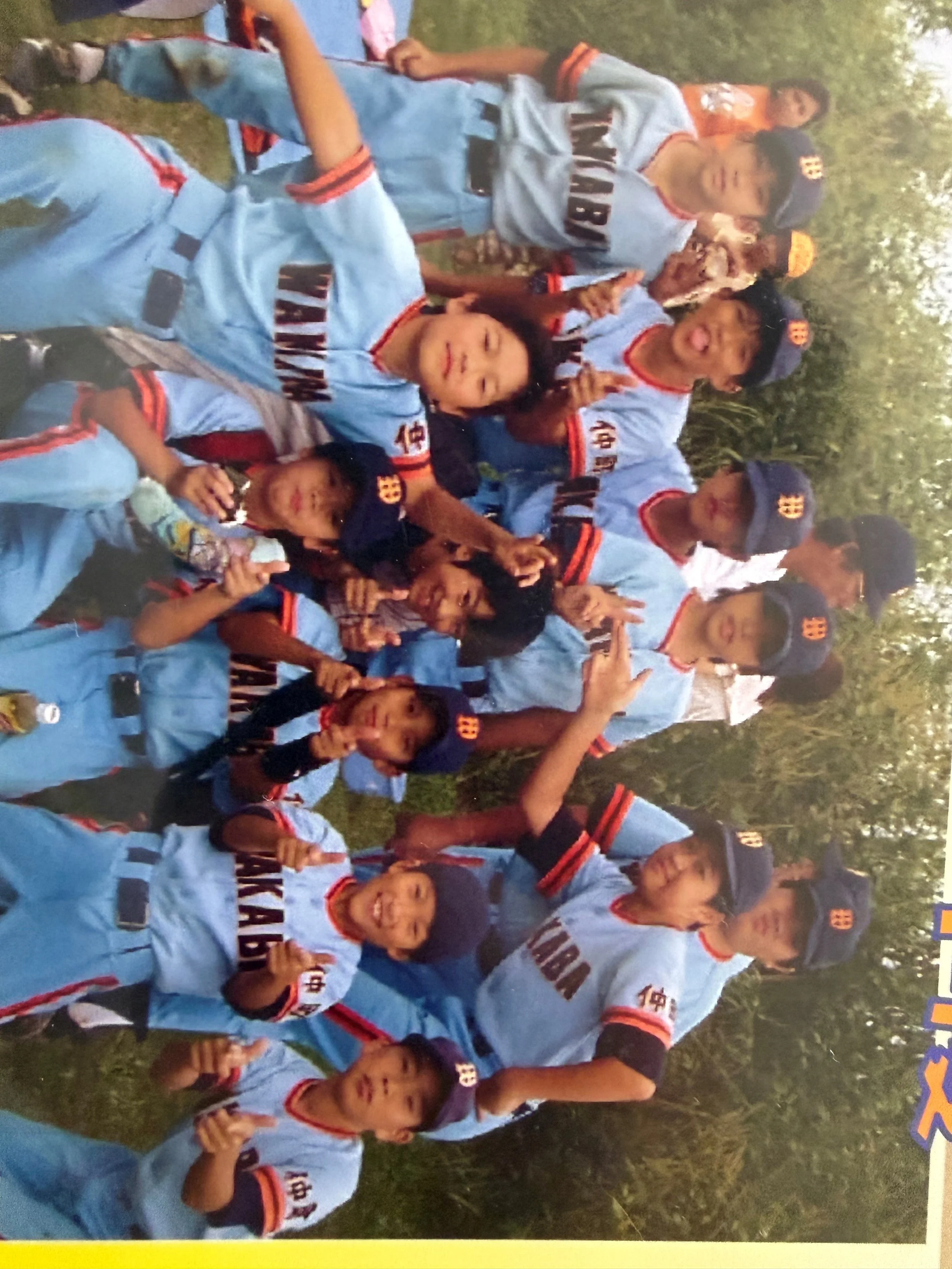 A group of children wearing matching blue sports uniforms and caps, some making peace signs, gathered outdoors on grass with trees in the background.