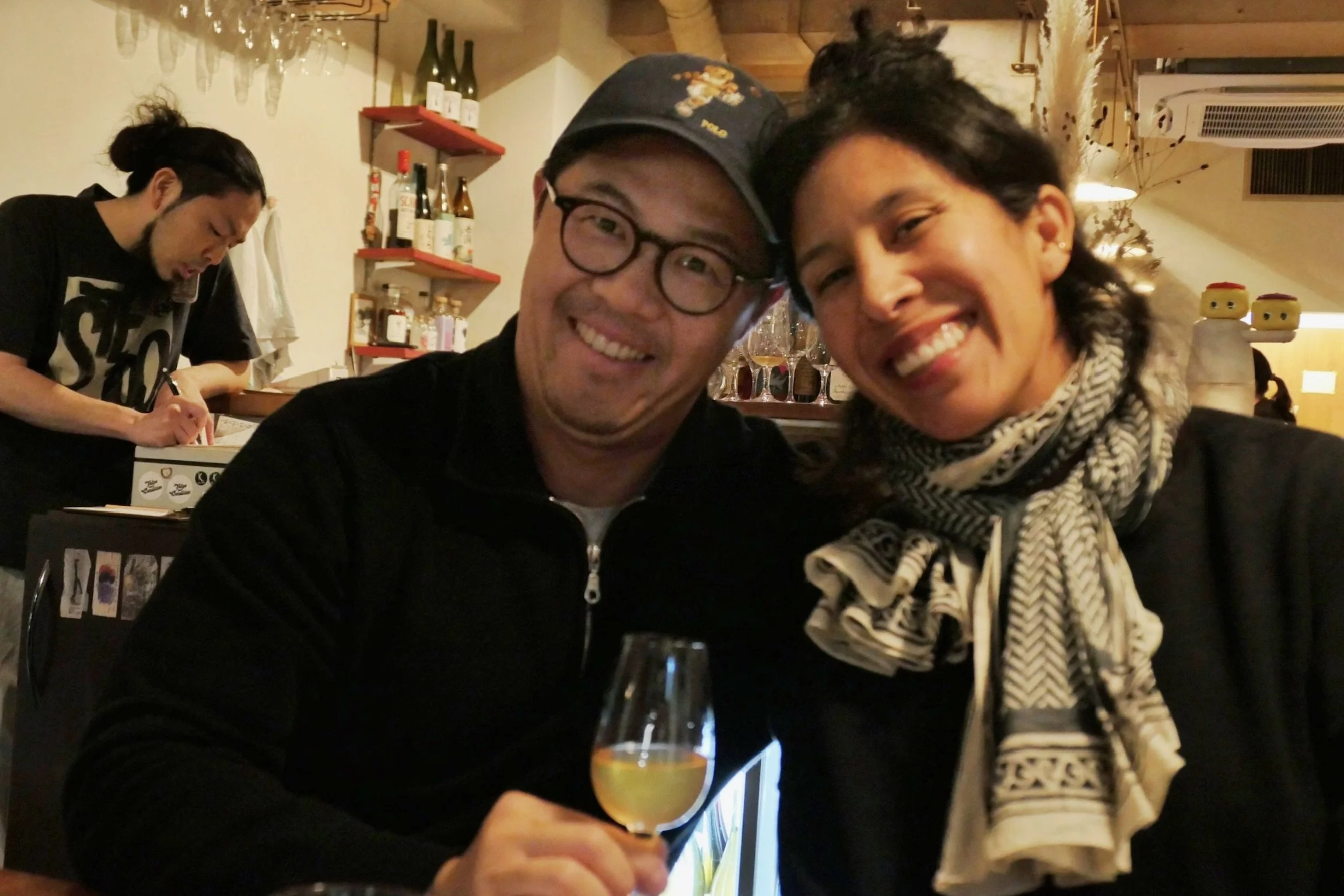 A man and woman smiling at the camera while sitting at a bar or restaurant, with a glass of drink in the foreground. There is a bartender in the background pouring a drink.