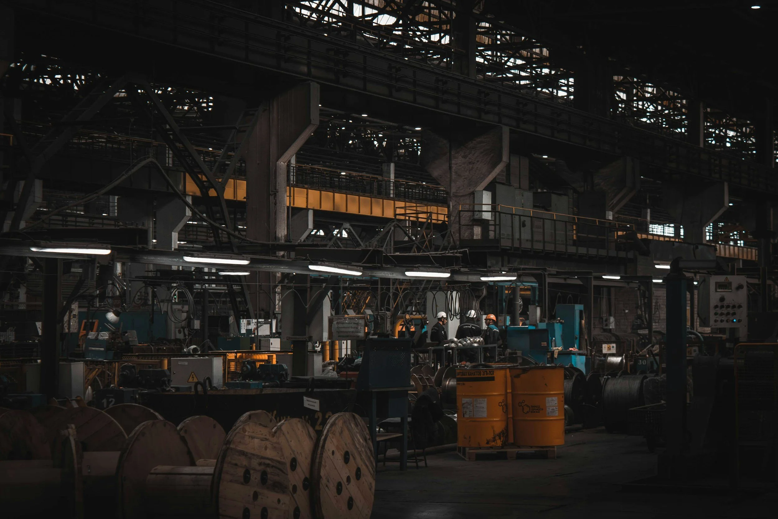 Inside an industrial factory with workers wearing safety helmets, large wooden spools, machinery, and overhead metal structures with visible lighting.