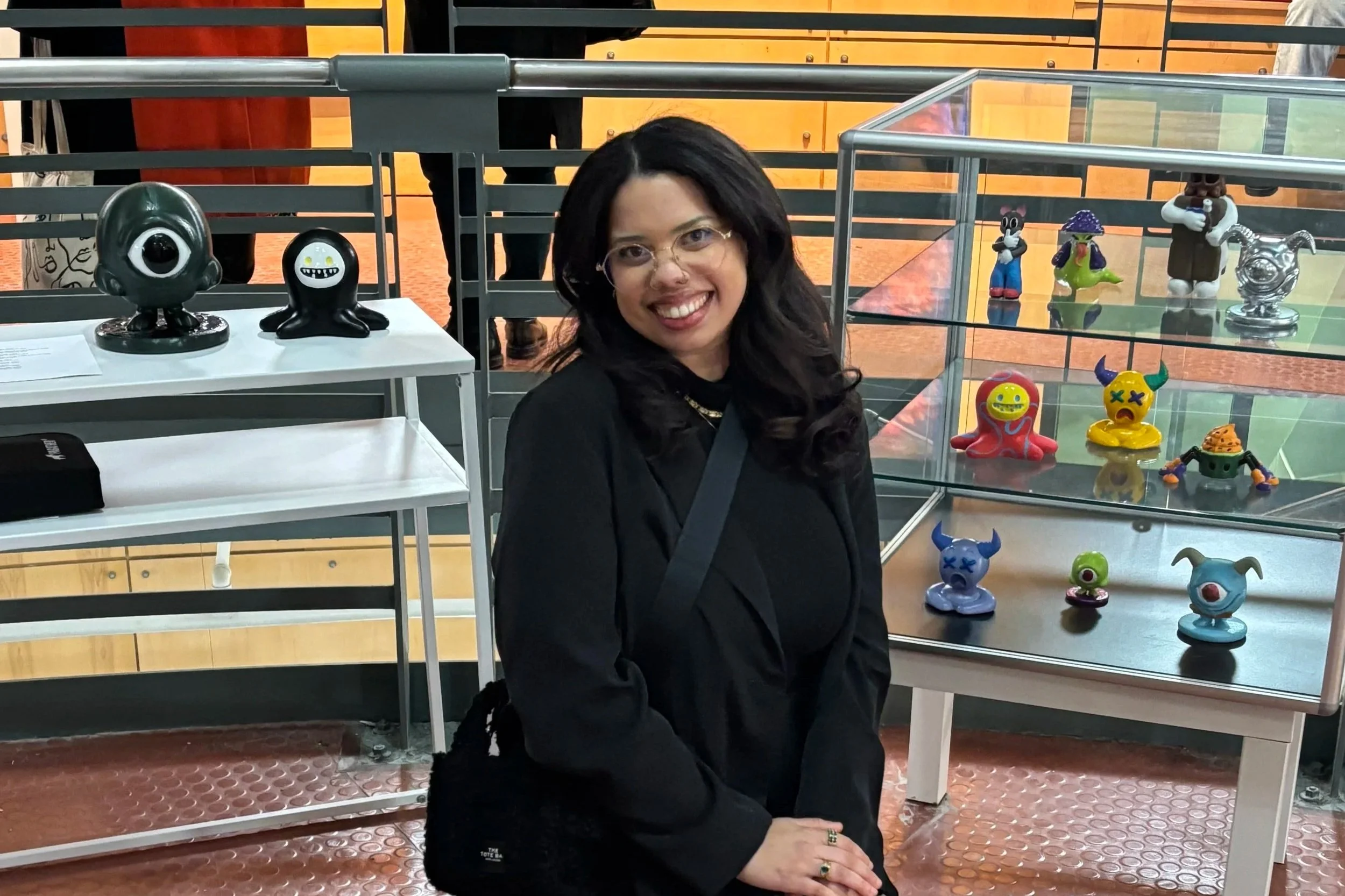 A woman with dark hair and glasses smiling while posing in front of glass display cases filled with colorful, quirky figurines and toys, at an indoor exhibition or gallery.
