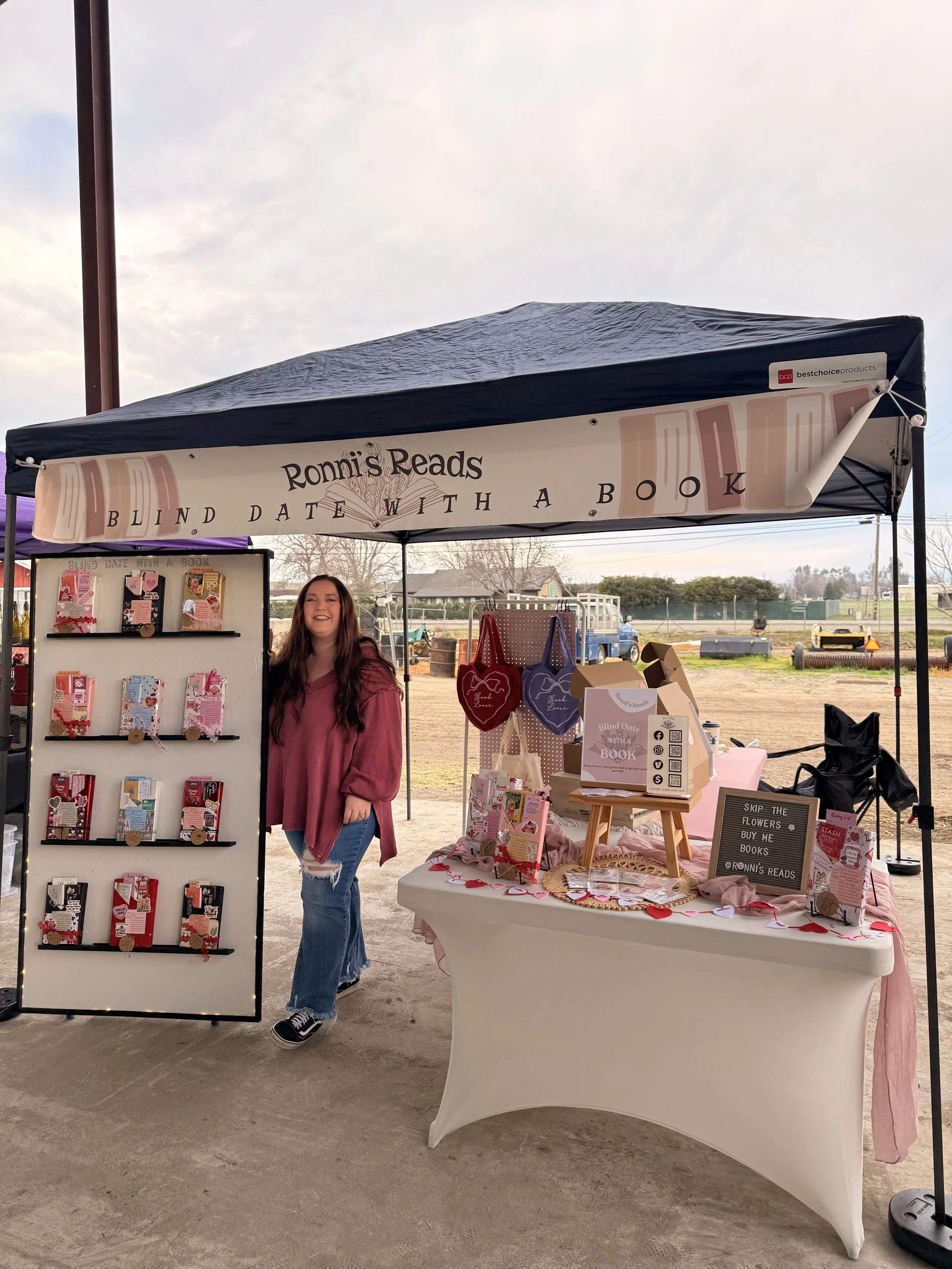 A woman standing behind a display booth at an outdoor market. The booth has a banner that reads "Ronni's Reads" and offers books for blind dates with a book. The table is decorated with hearts and pink decorations. Behind her, there are shelves with more books and a sign with information about the event.