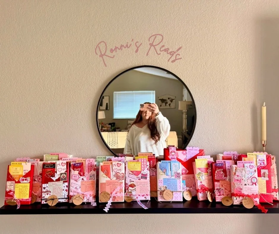 A mirror above a black shelf decorated with decorated book-themed paper bags, with a woman taking a selfie reflected in the mirror. The wall above the mirror has pink cursive writing that says "Romi's Reads."