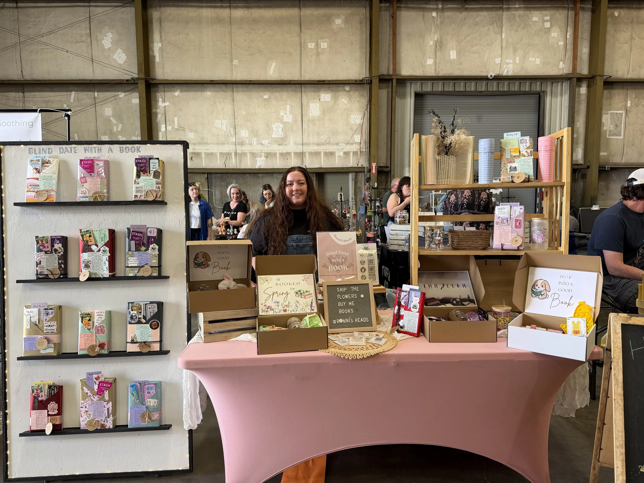 A woman at a book-themed craft booth, with shelves of bookmarks and a pink tablecloth, inside a warehouse-like space with exposed walls and ceiling.
