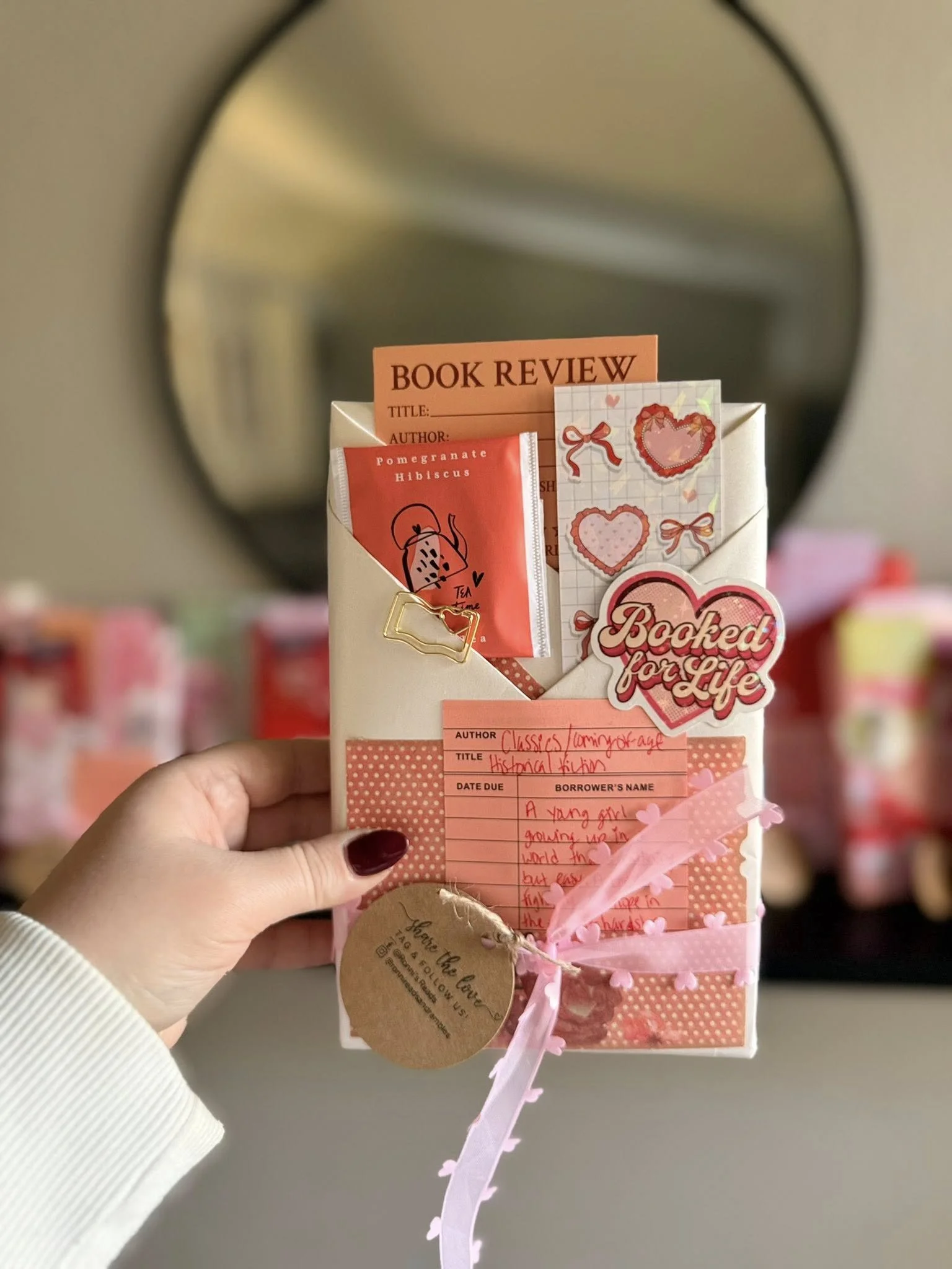 A person holding a pink and white decorated envelope with a tag that reads "Thanks for the love". The envelope contains a tea bag labeled "Pomegranate Hibiscus" and a small note with handwriting. There are various small paper hearts attached to the envelope and a sticker that says "Blocked for Life" in pink and white.