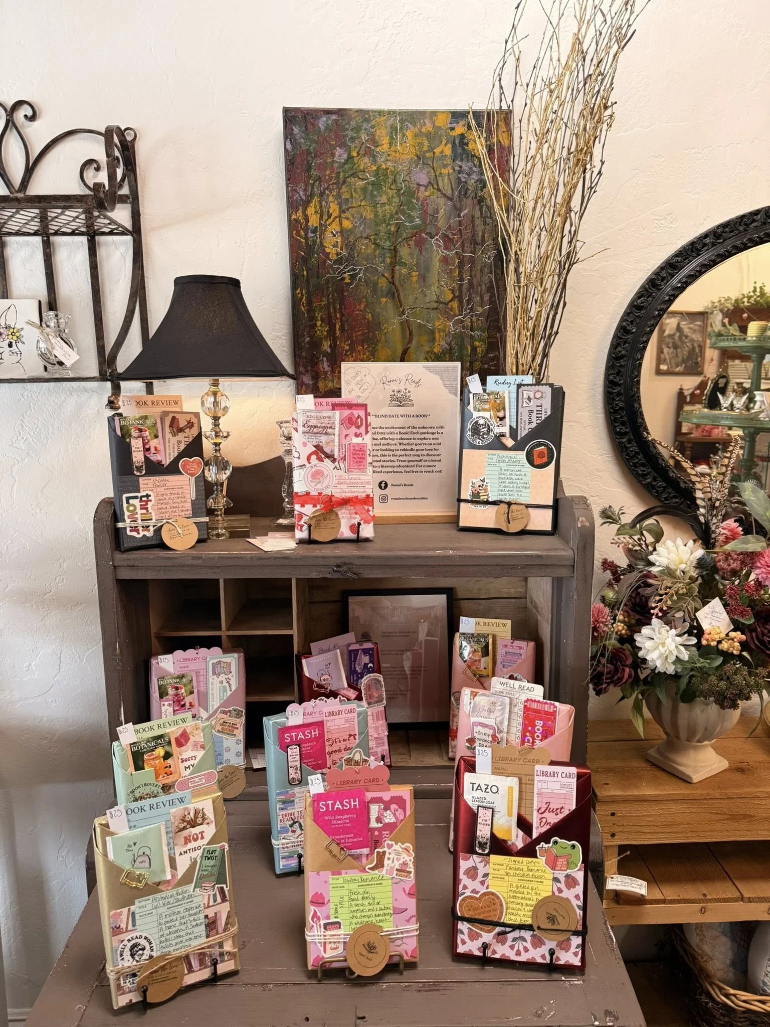 Display of various brightly colored notebooks, greeting cards, and paper crafts on a wooden table, with a large floral arrangement and a decorative mirror in the background.