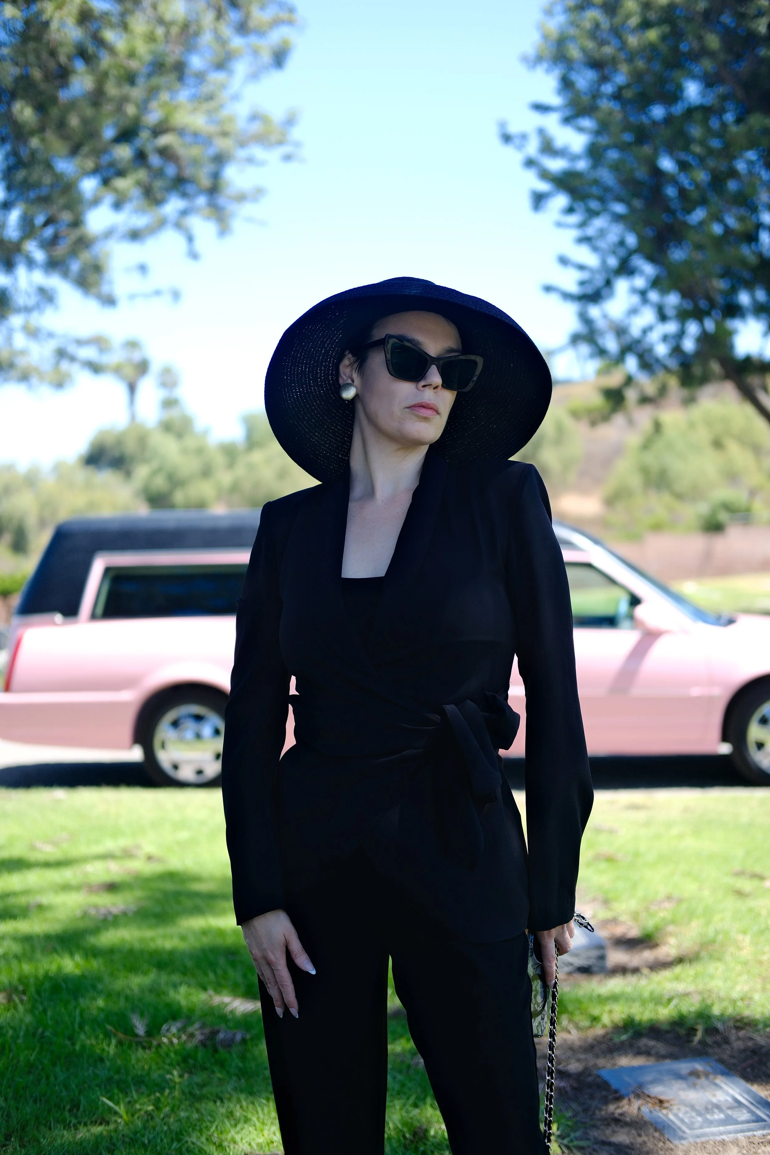 A woman wearing a black wide-brimmed hat, large sunglasses, pearl earrings, and a black outfit stands outdoors in front of a pink and white car with a background of green trees and blue sky.