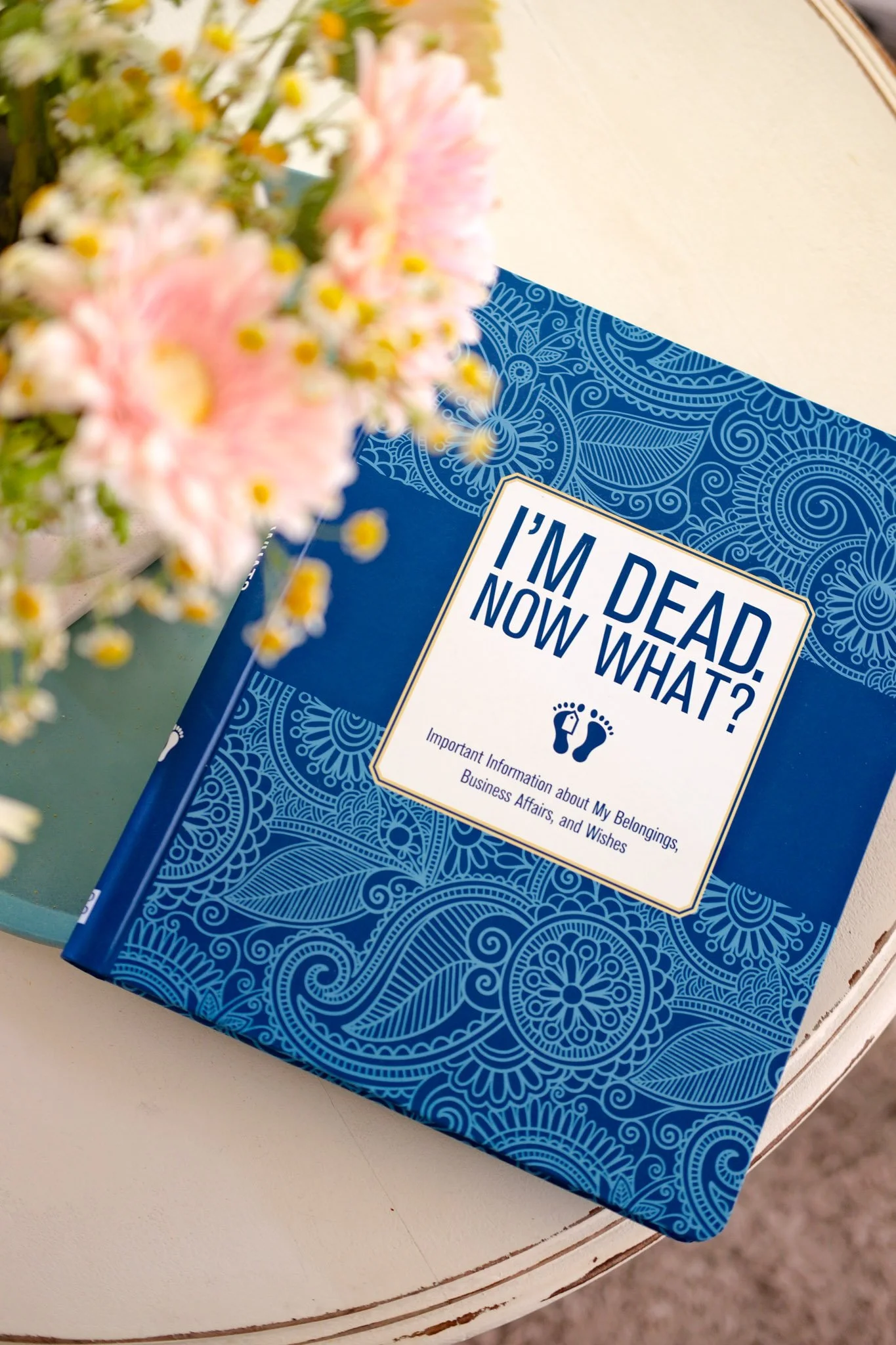 A blue book titled 'I'm Dead, Now What?' is placed on a white round table. A vase of pink and white flowers is in the foreground, partially obscuring the book.