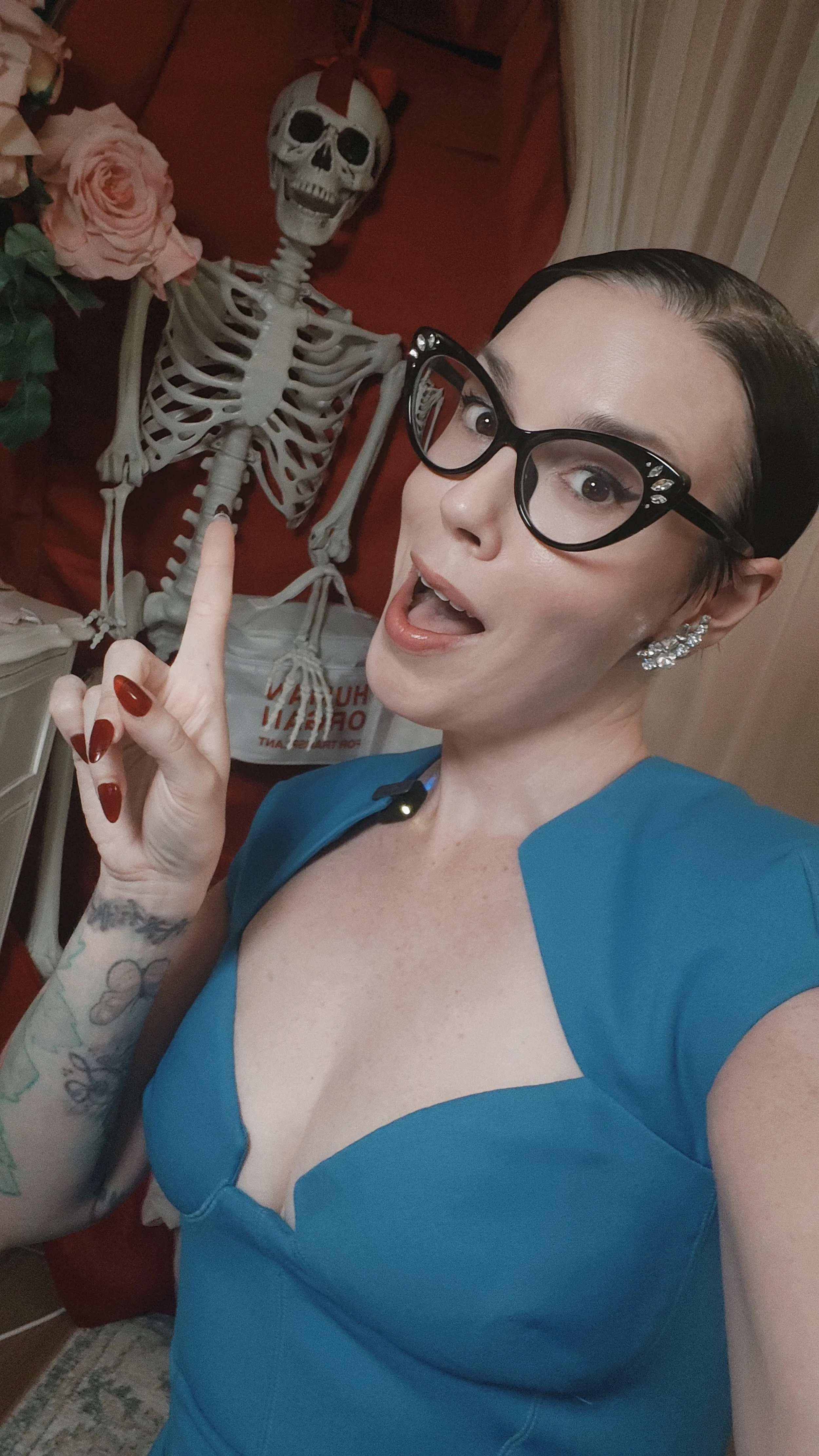 A woman wearing glasses and a blue top posing for a selfie with a skeleton decorated with a red ribbon around its skull and a pink rose on its shoulder in the background.