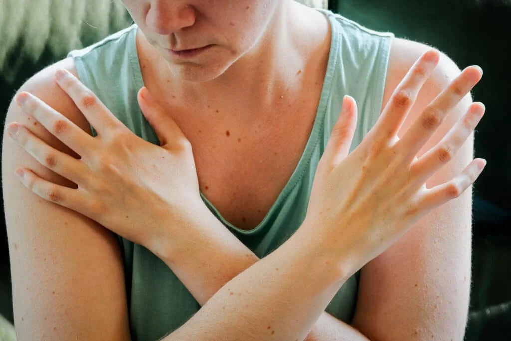 A woman with crossed arms, wearing a sleeveless green top, is holding her shoulders with her hands. The image is a close-up of her upper body. This bi-lateral movement helps with regulating our emotions.