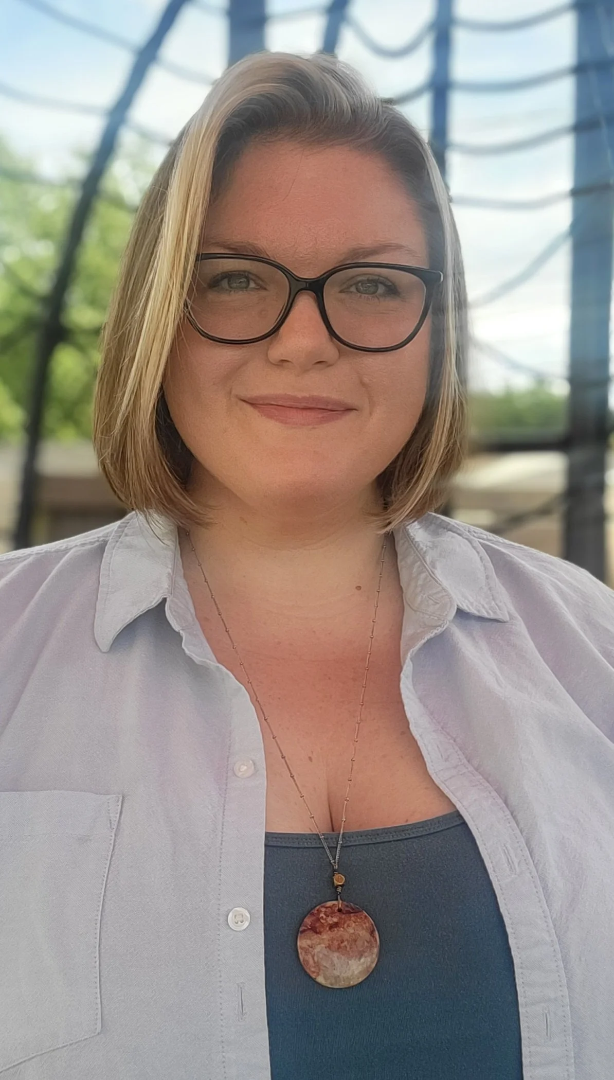 A woman with shoulder-length blonde hair and glasses, wearing a necklace with a round pendant, a dark top, and a light shirt, standing outdoors with a blurred background of trees and metal structures.