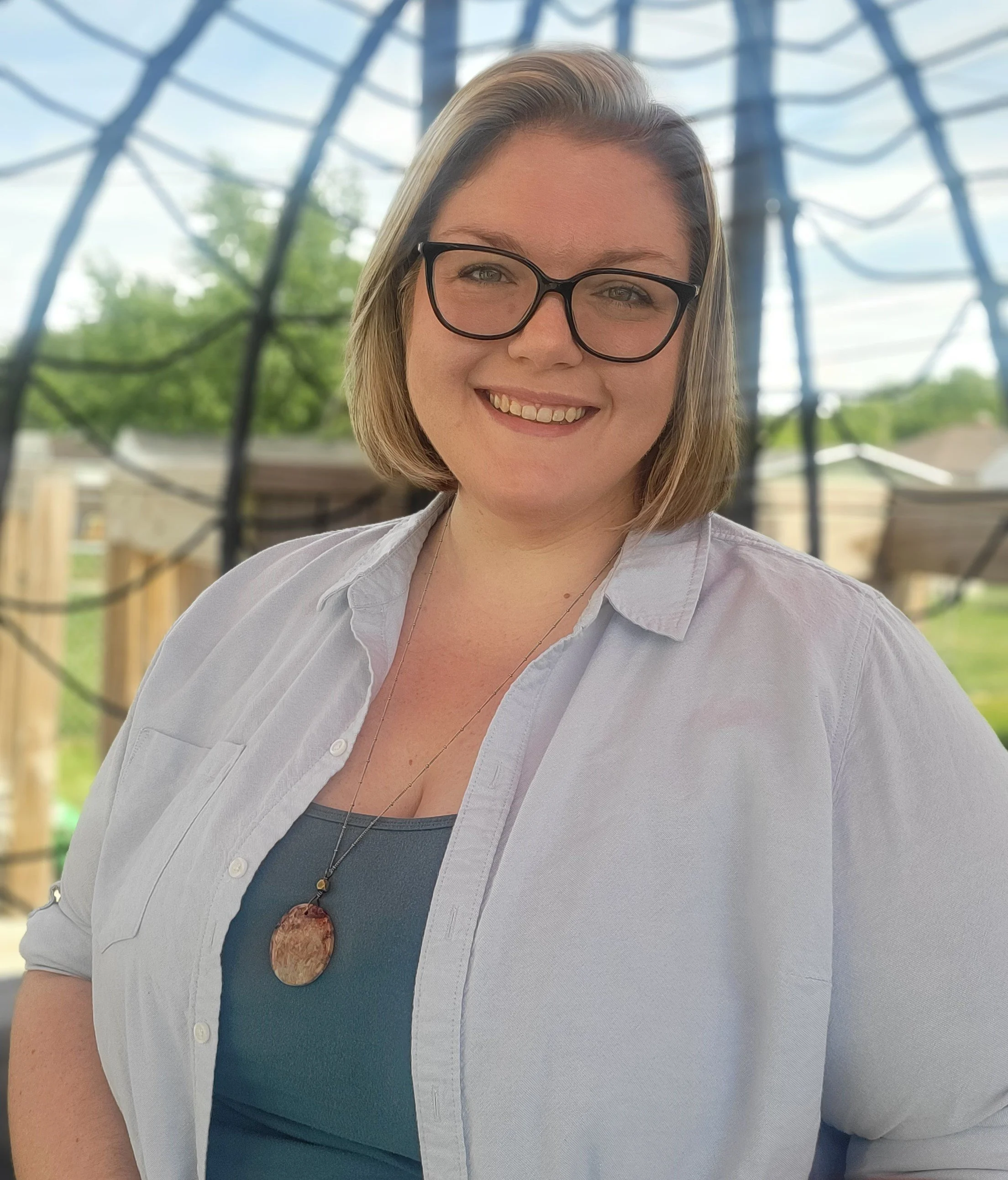 A woman with blonde hair, glasses, and a gray shirt, smiling outdoors with a blurred background of trees and a structure.