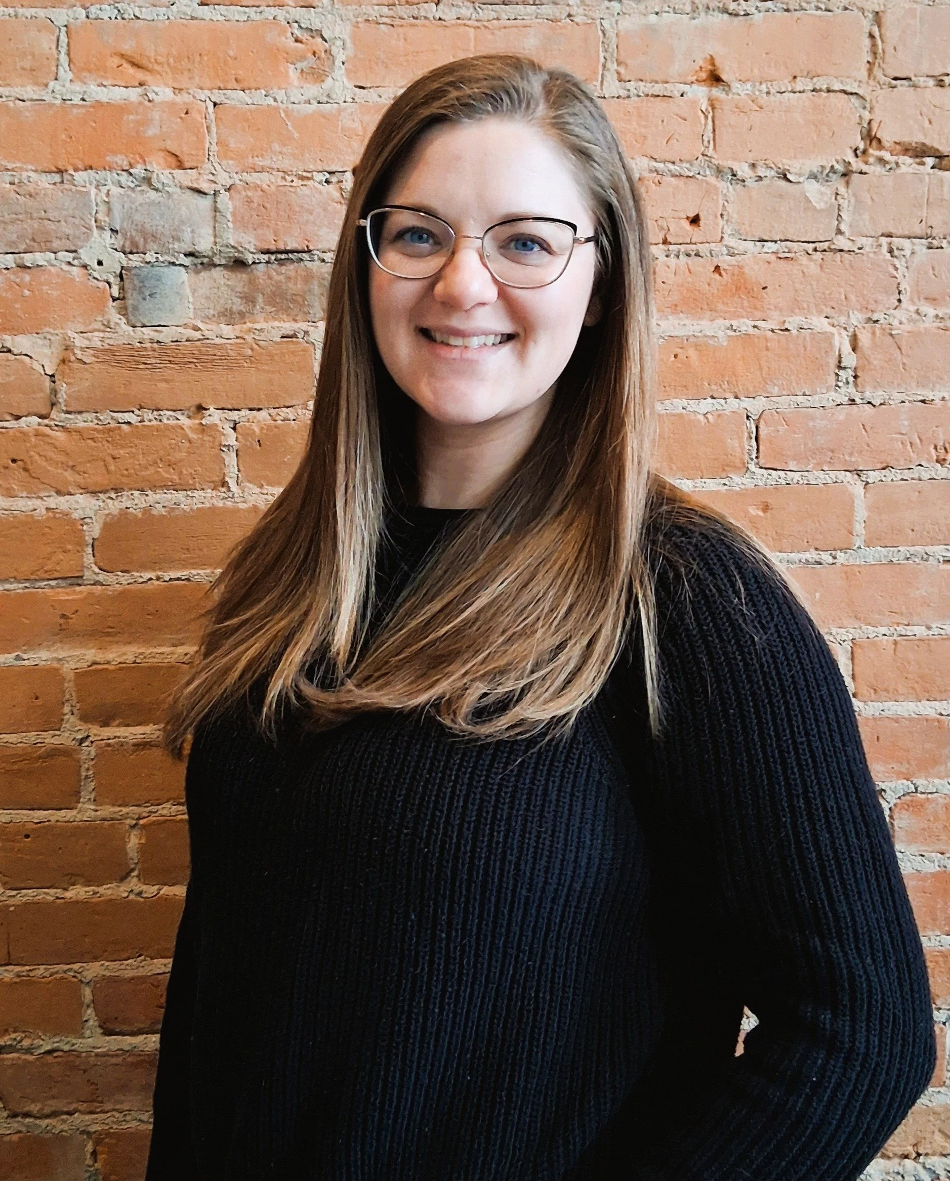 A smiling woman with long blonde hair and glasses in front of a red brick wall.