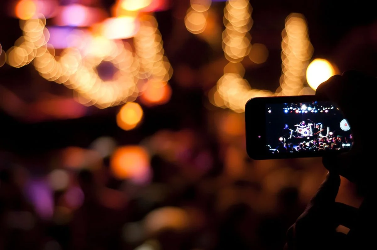 A person using a smartphone to record a live concert with a band performing on stage, surrounded by blurred colorful lights.