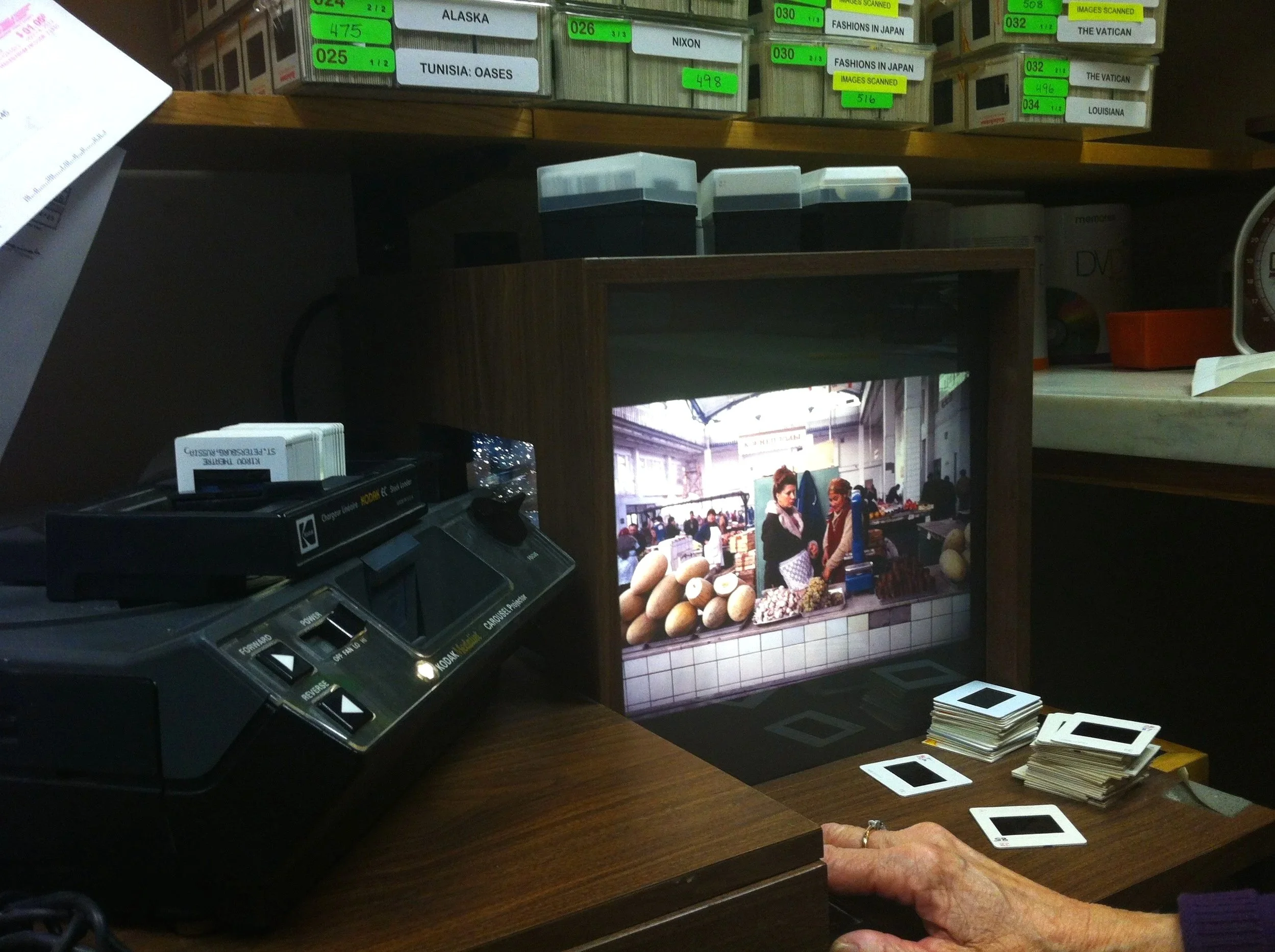 A slide viewer box on a desk and the user's hand. There are stacks of color slides on the desk and shelves above with labeled boxes containing various items.