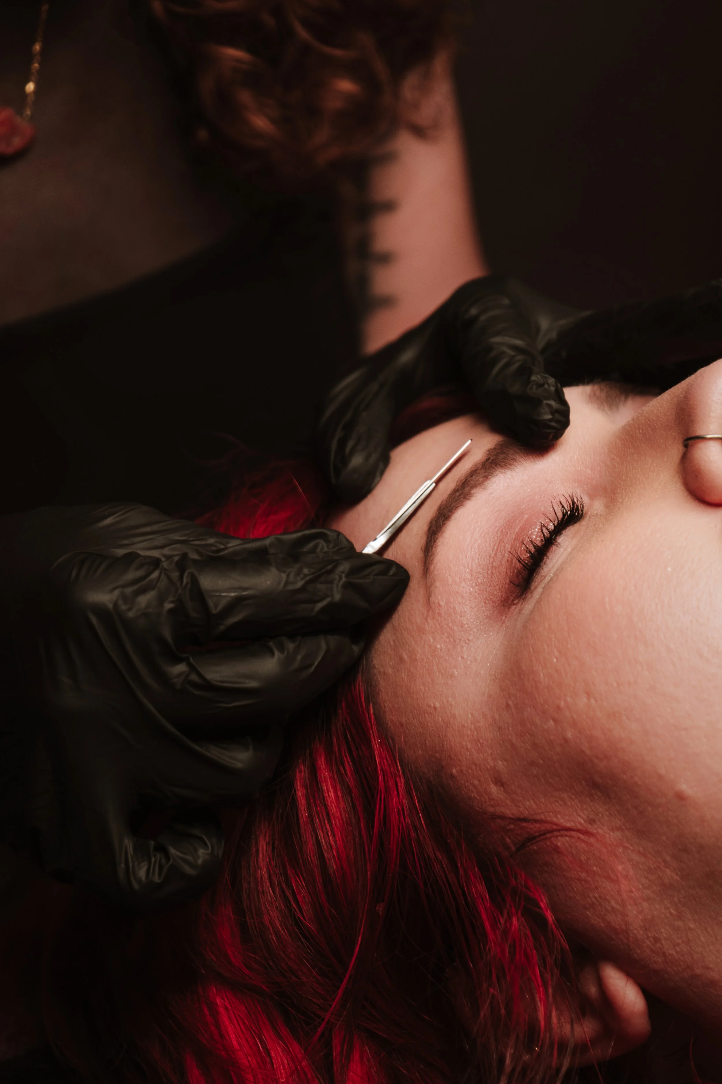 Cosmetologist performs a facial piercing on a young woman with red hair, using a needle while wearing black gloves.