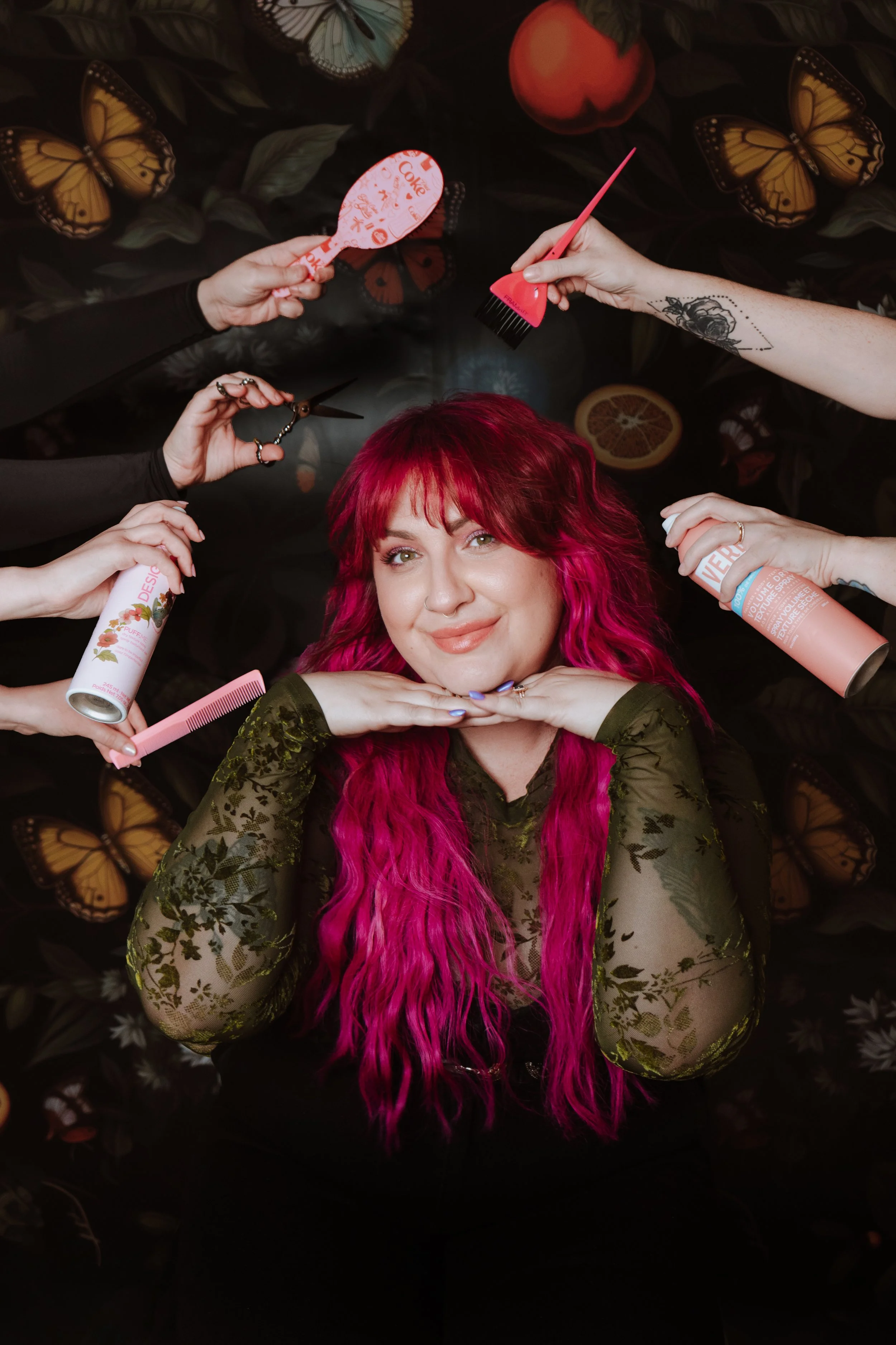 A woman with vibrant pink hair lying on a dark floral background, surrounded by hands holding various beauty and haircare products, with colorful butterflies and dried citrus slices in the background.
