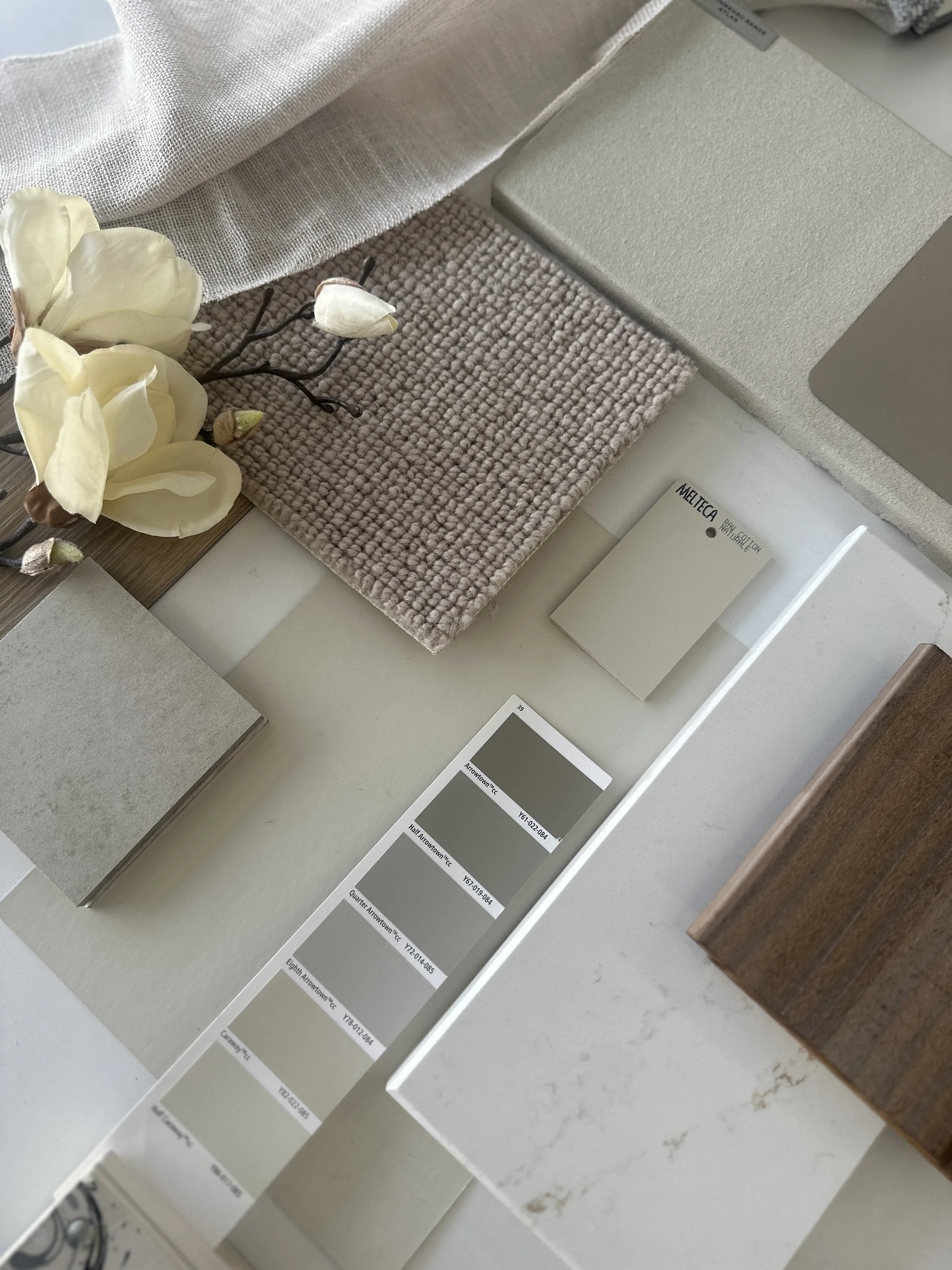 Swatches of paint colors on color sample cards with labels, a beige textured fabric, a white textured material, a small beige carpet sample, a white book, and artificial white flowers with dark branches.