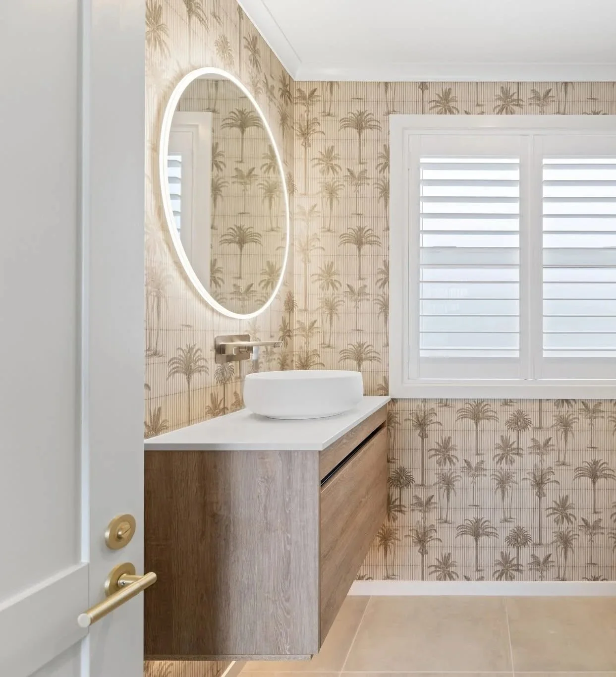 Bathroom with palm tree wallpaper, a wall-mounted sink, illuminated mirror, and a window with white shutters.