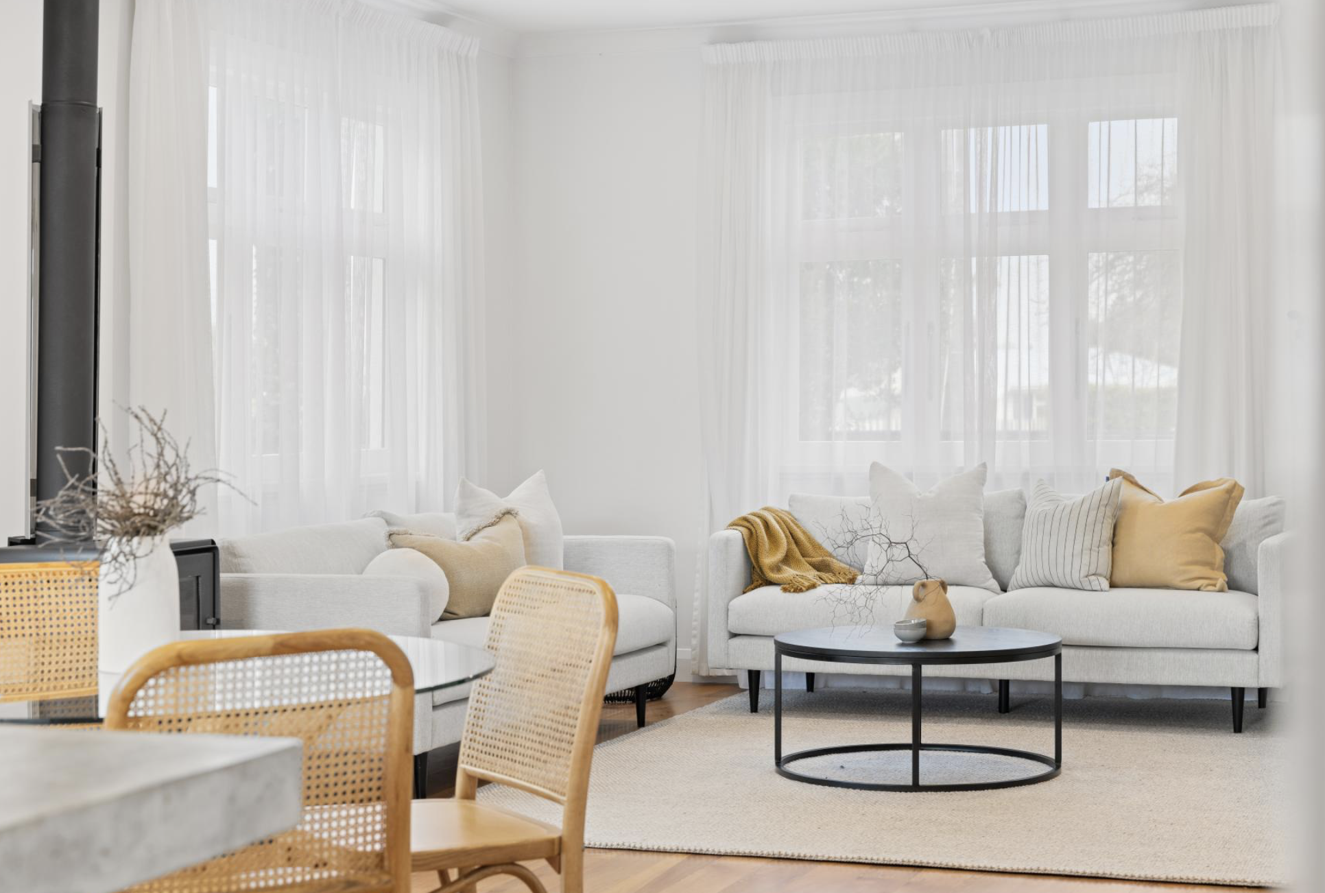 Bright living room with large windows, white curtains, white sofa, beige and yellow cushions, black coffee table with decorative objects, and a textured beige rug. Part of a dining area with wooden chairs is visible in the foreground.