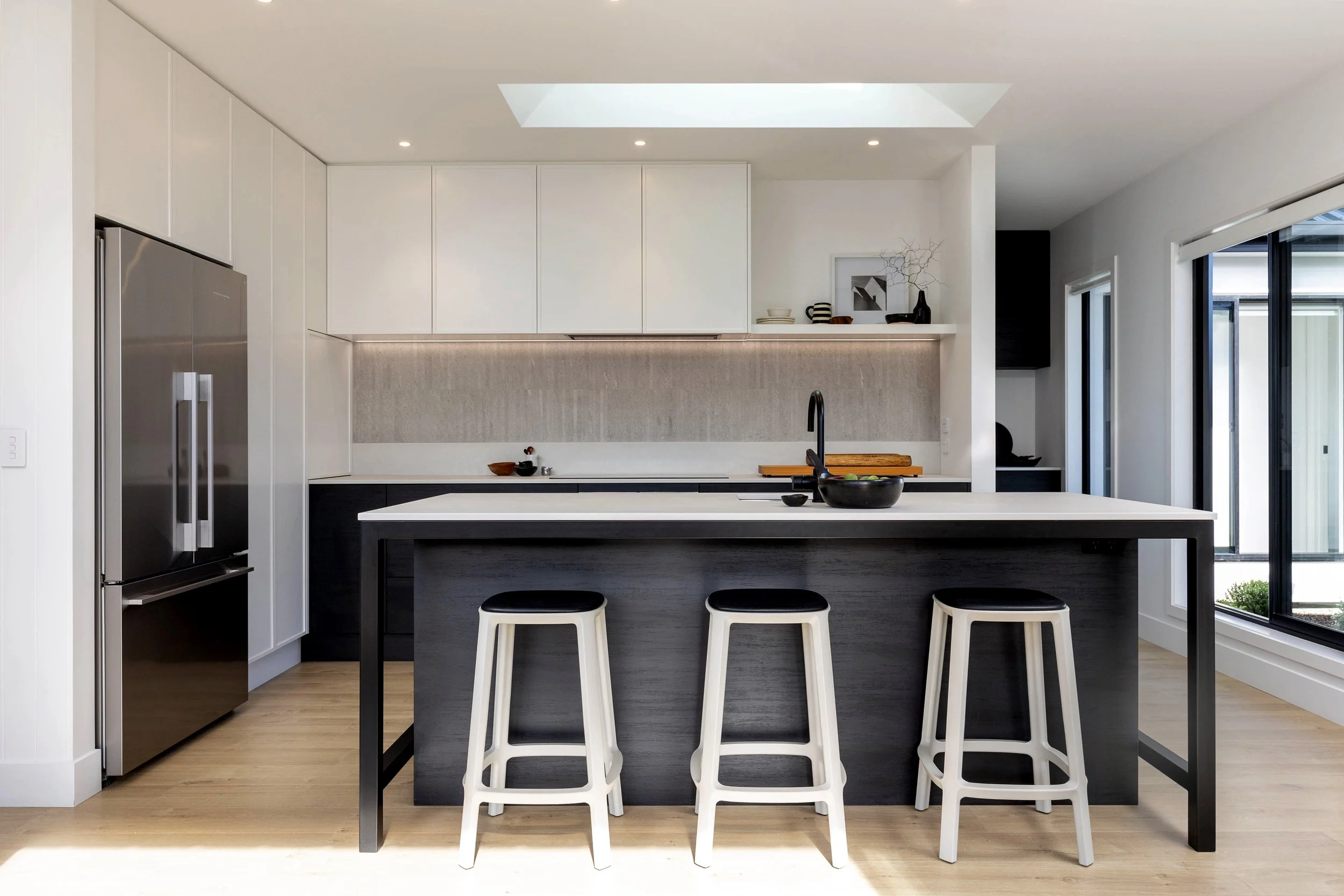 Modern kitchen with white cabinets, black island with bar stools, stainless steel refrigerator, and large windows with views of the outdoors.