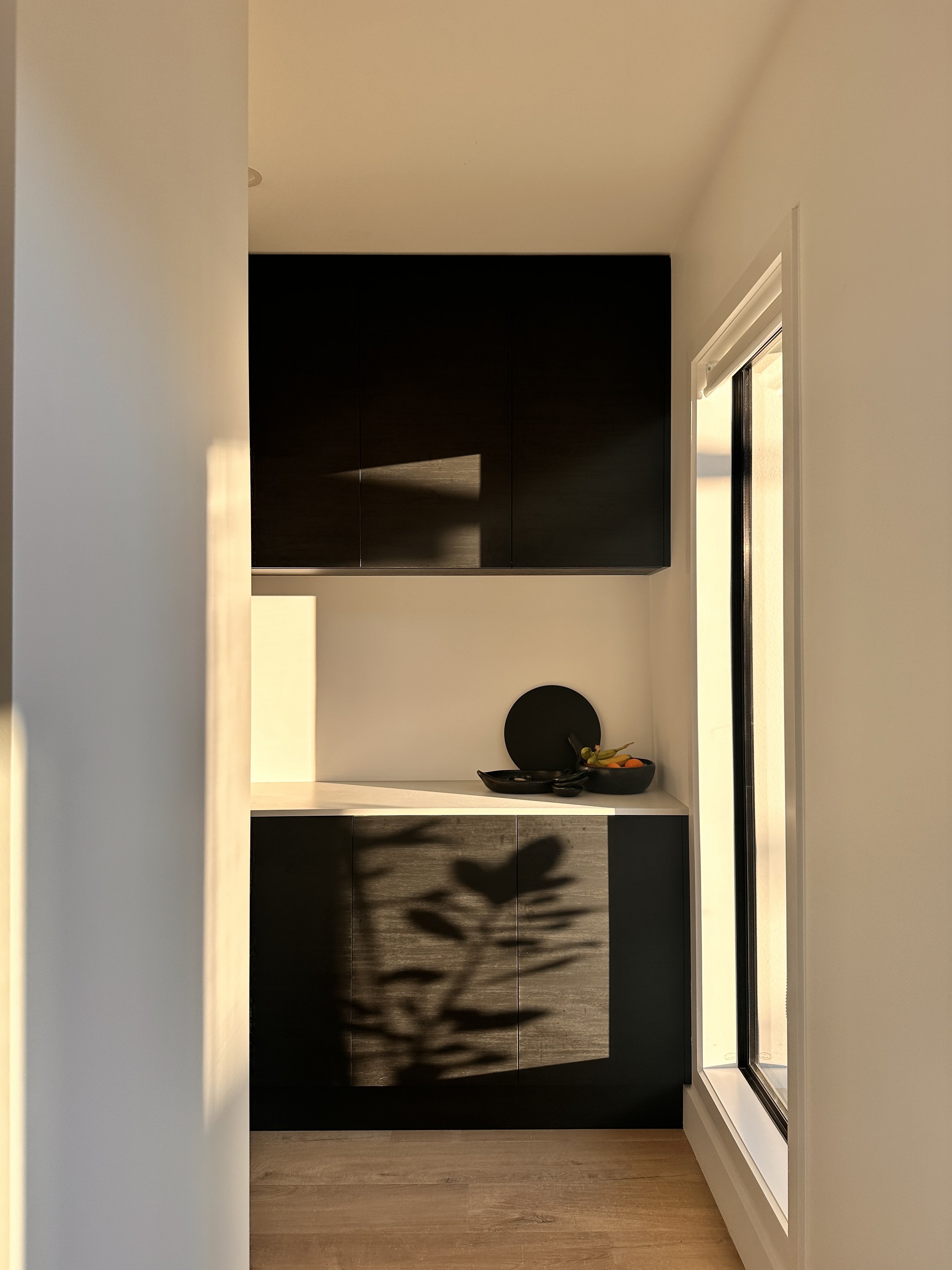 A modern kitchen with black cabinets, a white countertop, a bowl of fruit, and a large window allowing natural light.