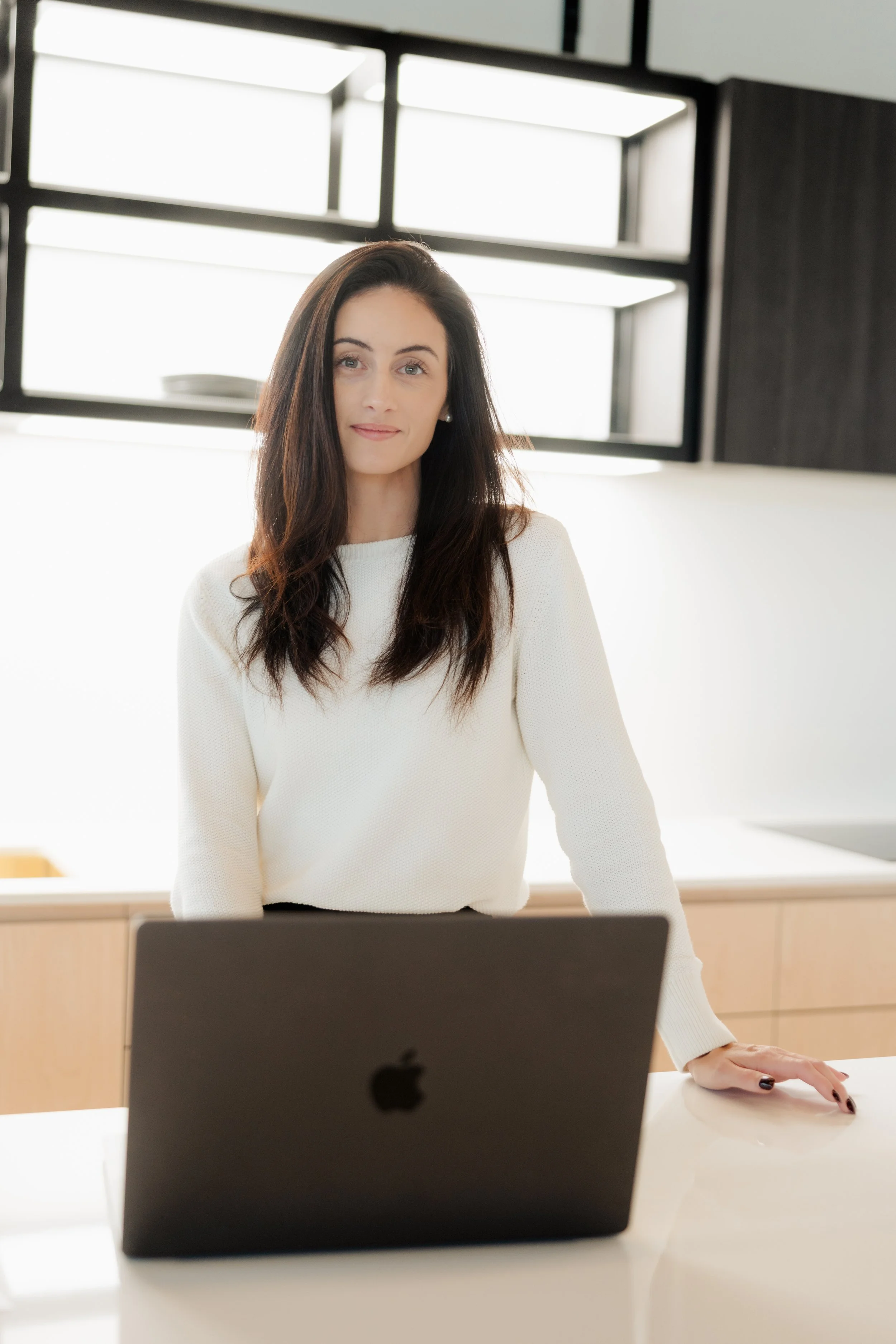 A woman with long dark hair and a cream-colored sweater sitting at a desk in a bright office with modern decor, with a laptop in front of her.