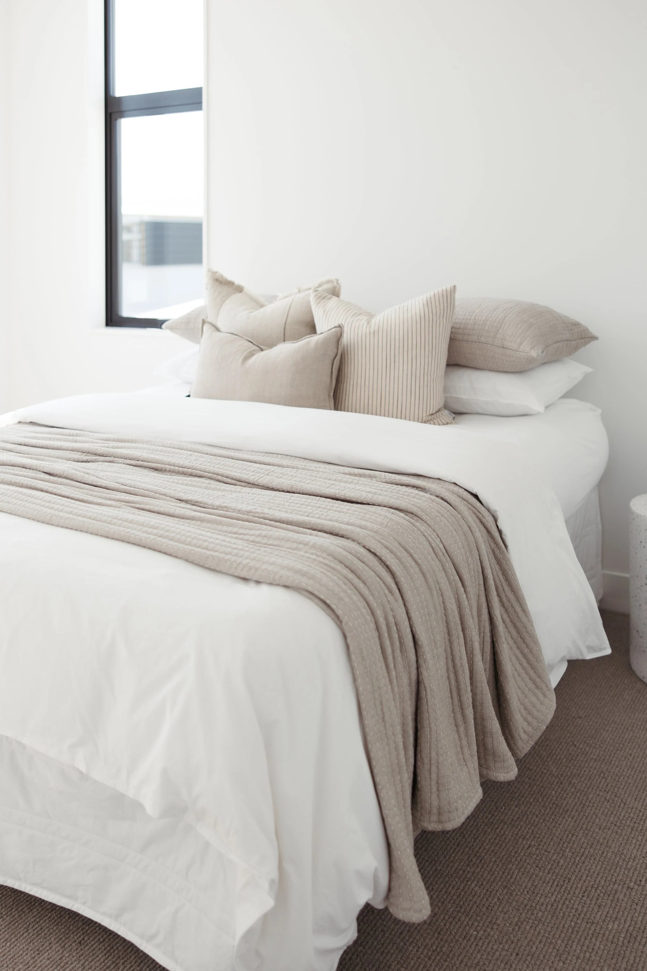 A neatly made bed with a white duvet cover and a beige textured blanket draped over the foot of the bed. Several beige and striped pillows are arranged on top, and there is a window with black framing on the left, allowing natural light into the room.