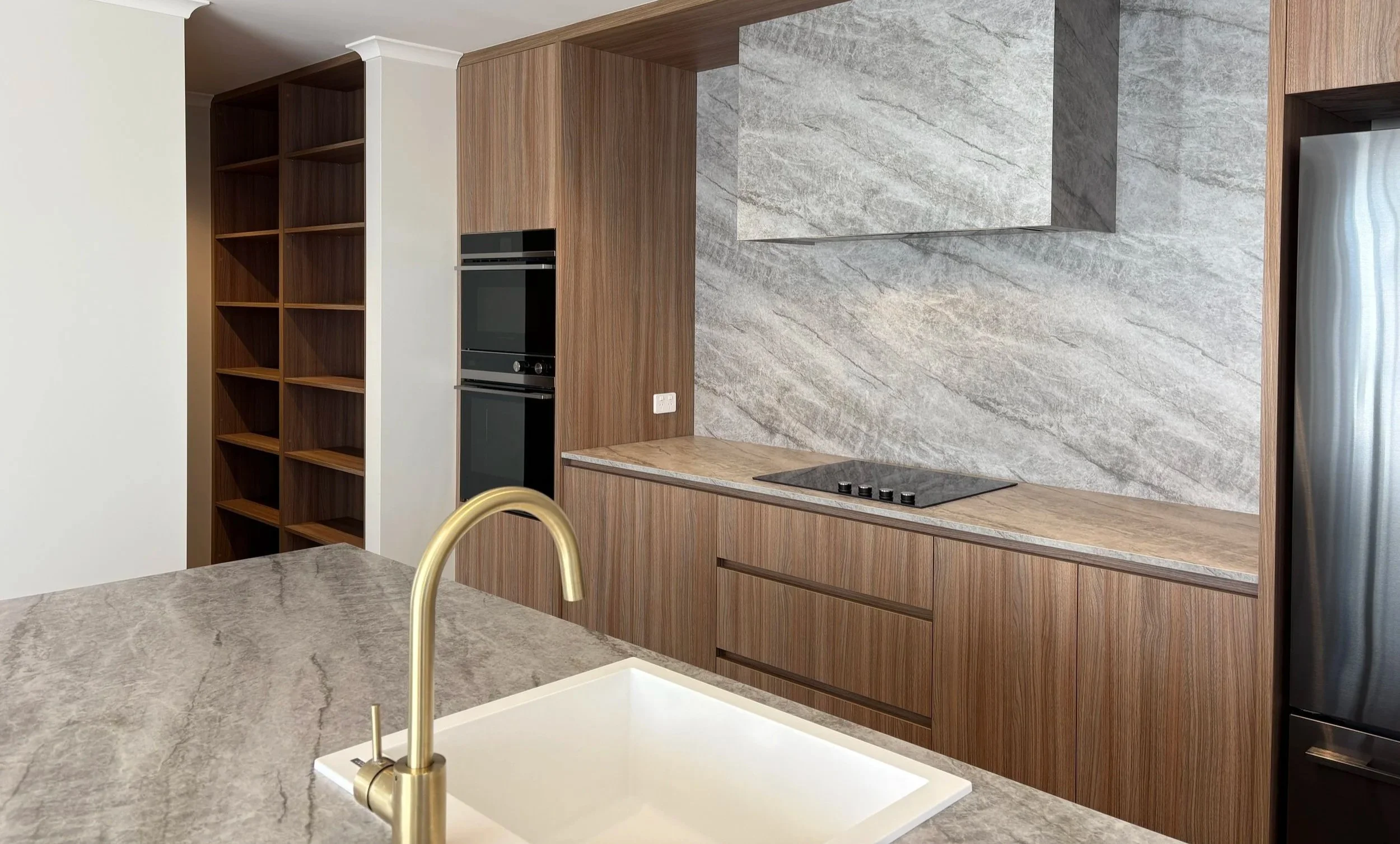 Modern kitchen with beige marble countertops, a built-in stovetop, wooden cabinetry, a marble backsplash, a dual oven, a stainless steel refrigerator, and a faucet over a white sink.