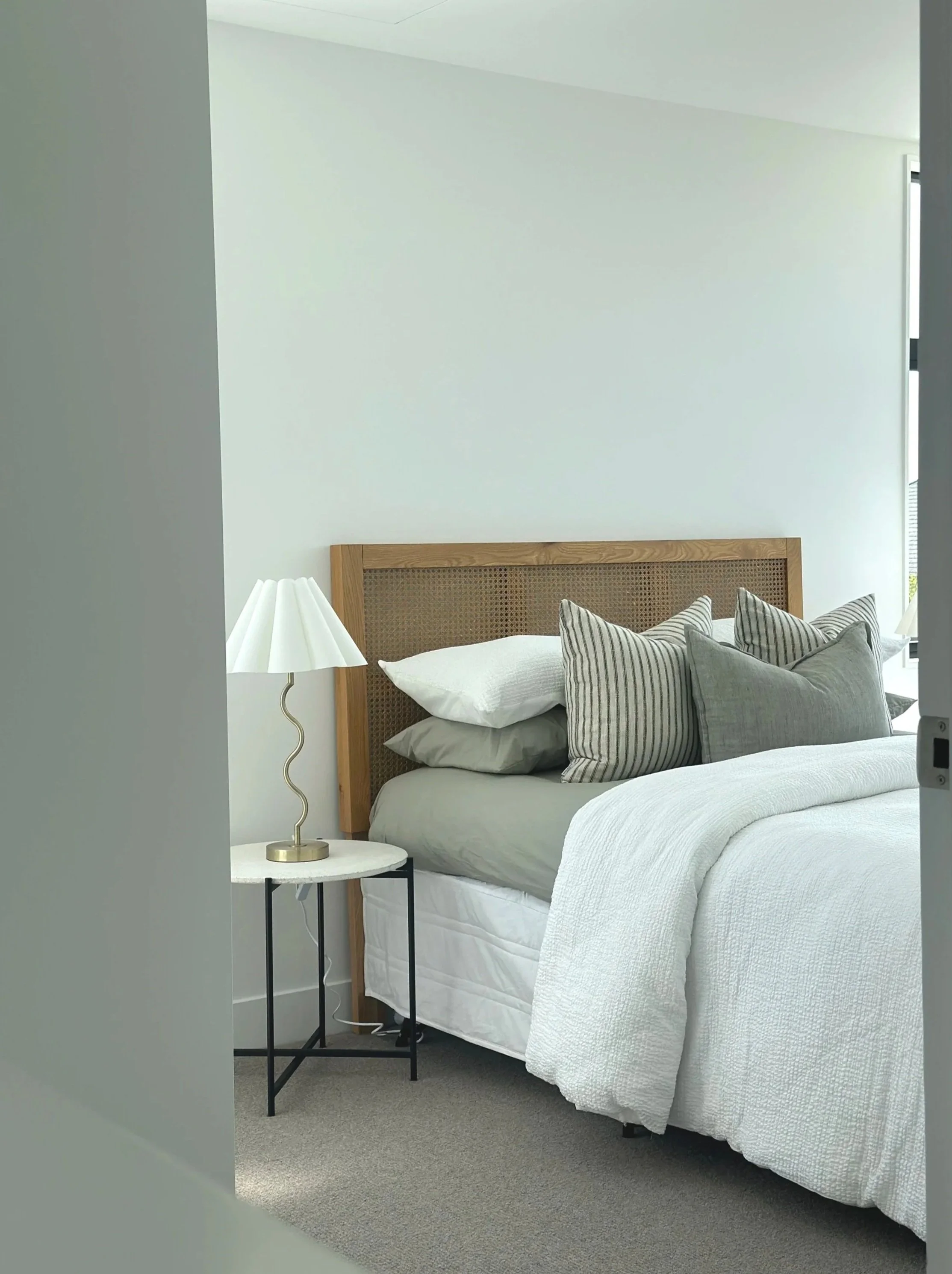 A bedroom with a bed, a headboard made of wood with woven cane, several pillows, a white quilt, and a small round side table with a modern white table lamp. There is a window in the corner of the room.