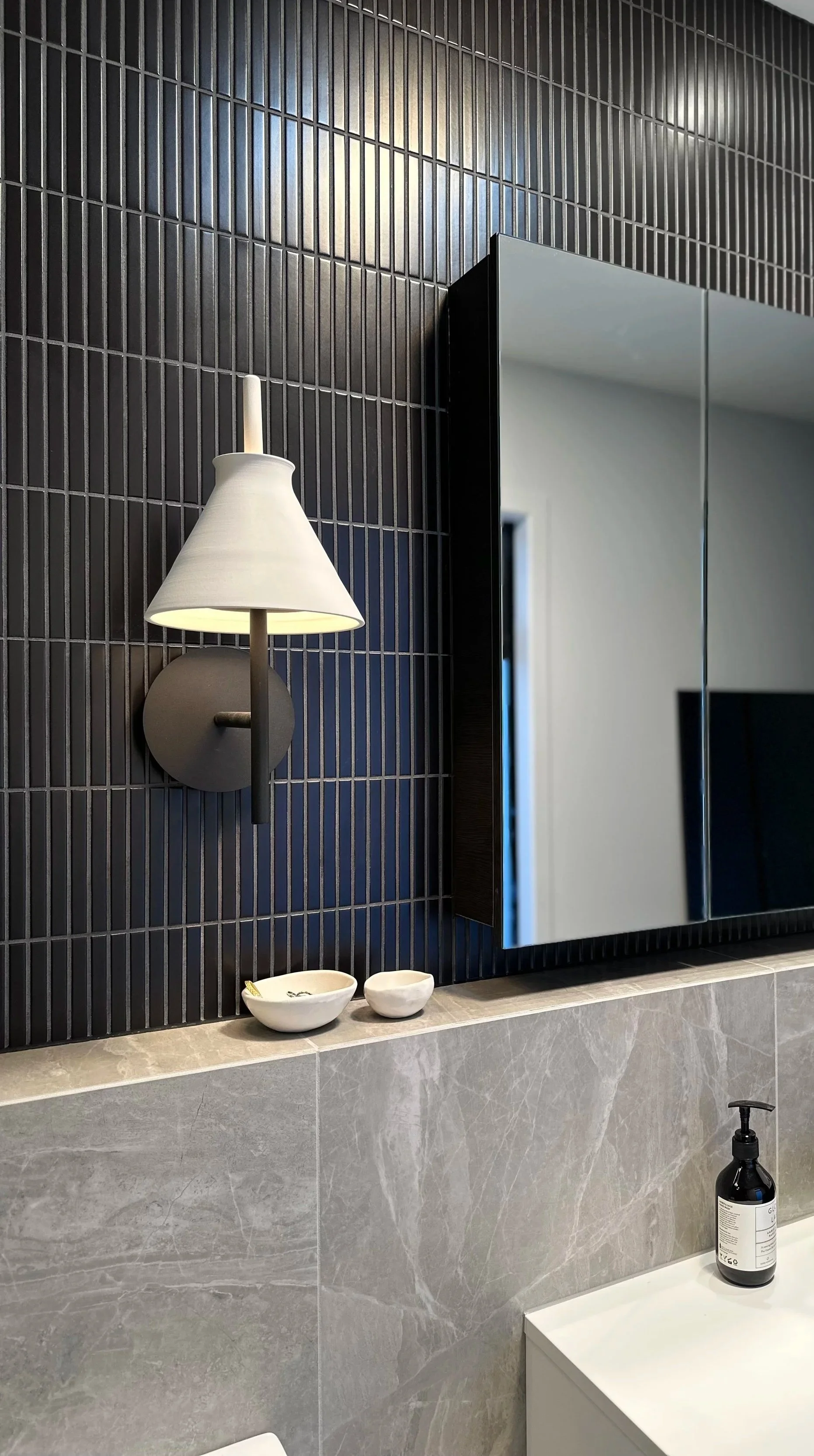 Modern bathroom with black tiled wall, a wall-mounted lamp, a mirror, and small ceramic bowls on the gray marble countertop. There is a black soap dispenser on a white surface below.