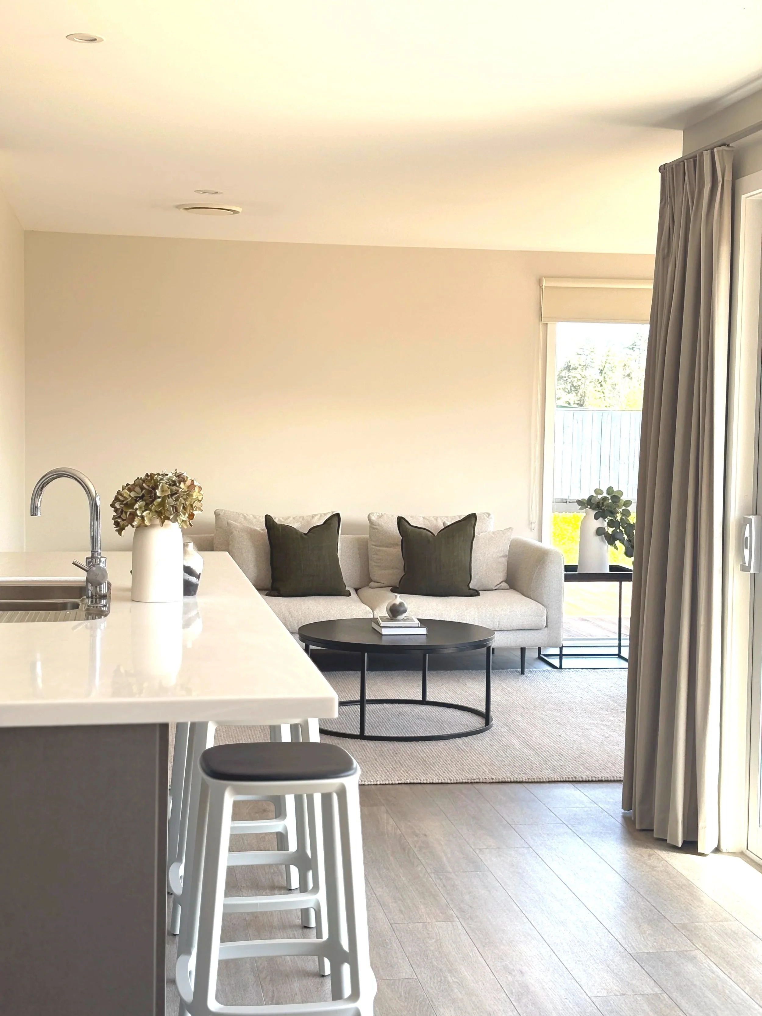 A bright, modern living room with a beige couch, black pillows, coffee table, and large windows with curtains, viewed from a kitchen island with white stools.